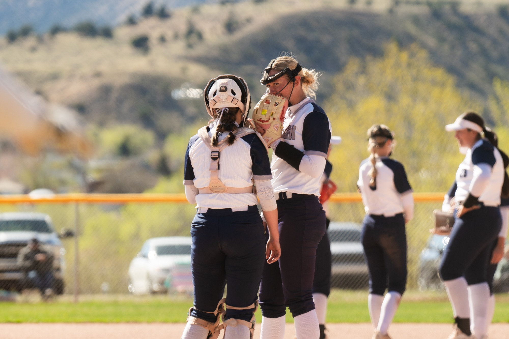 A candid, sunlit photo of the Utah State Eastern softball team on the field. A pitcher and catcher in white and navy uniforms are seen in a close conversation, with the pitcher covering her mouth with her glove. Other teammates are visible in the background against a backdrop of rolling hills and a clear sky.
