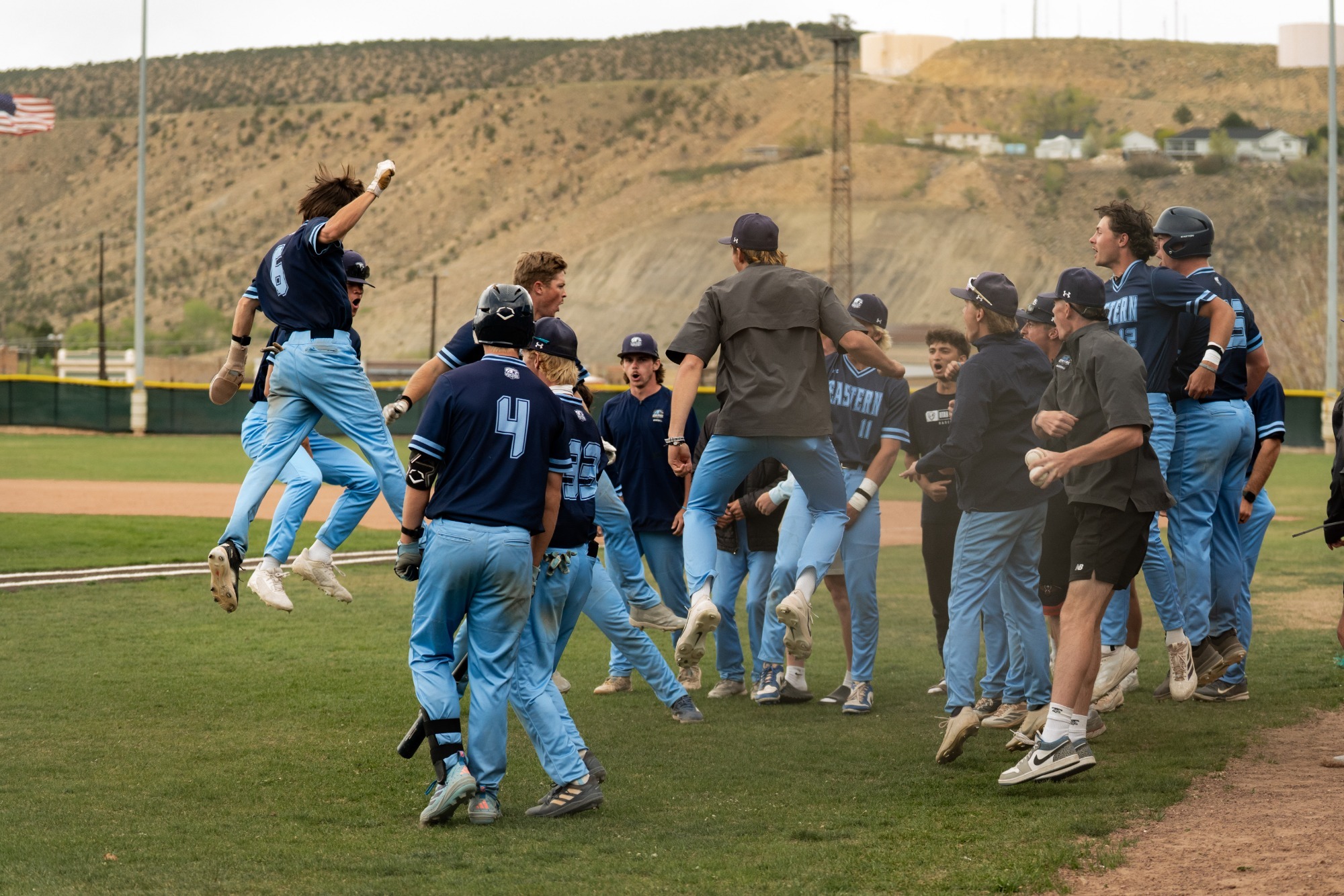 A high-energy, candid photo of the Utah State Eastern baseball team celebrating on the field. Several players in light blue and navy jerseys are captured mid-air jumping for joy, while others huddle together cheering. The scene is set on a green baseball diamond with desert hills and a few buildings in the background under an overcast sky.