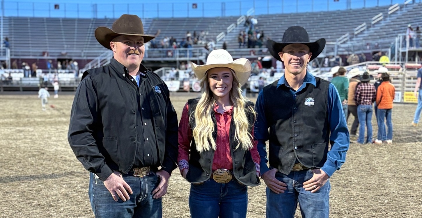 Three members of the Utah State Eastern rodeo team standing together in a dirt arena. They are wearing western attire, including cowboy hats, button-down shirts, vests, and jeans with large trophy belt buckles. An empty grandstand and a clear blue sky are in the background.