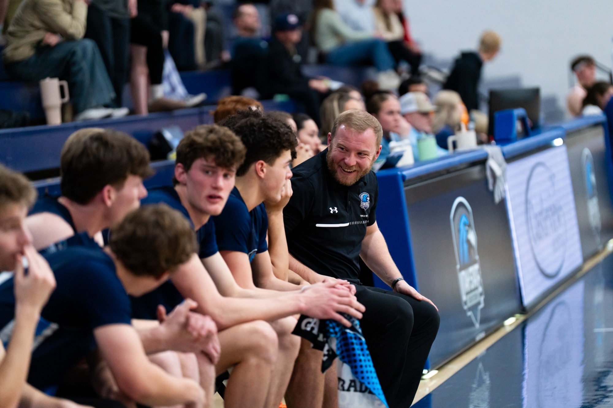 A basketball coach sits courtside smiling and talking with players on the bench during a game, while teammates listen and spectators fill the blurred bleachers in the background.