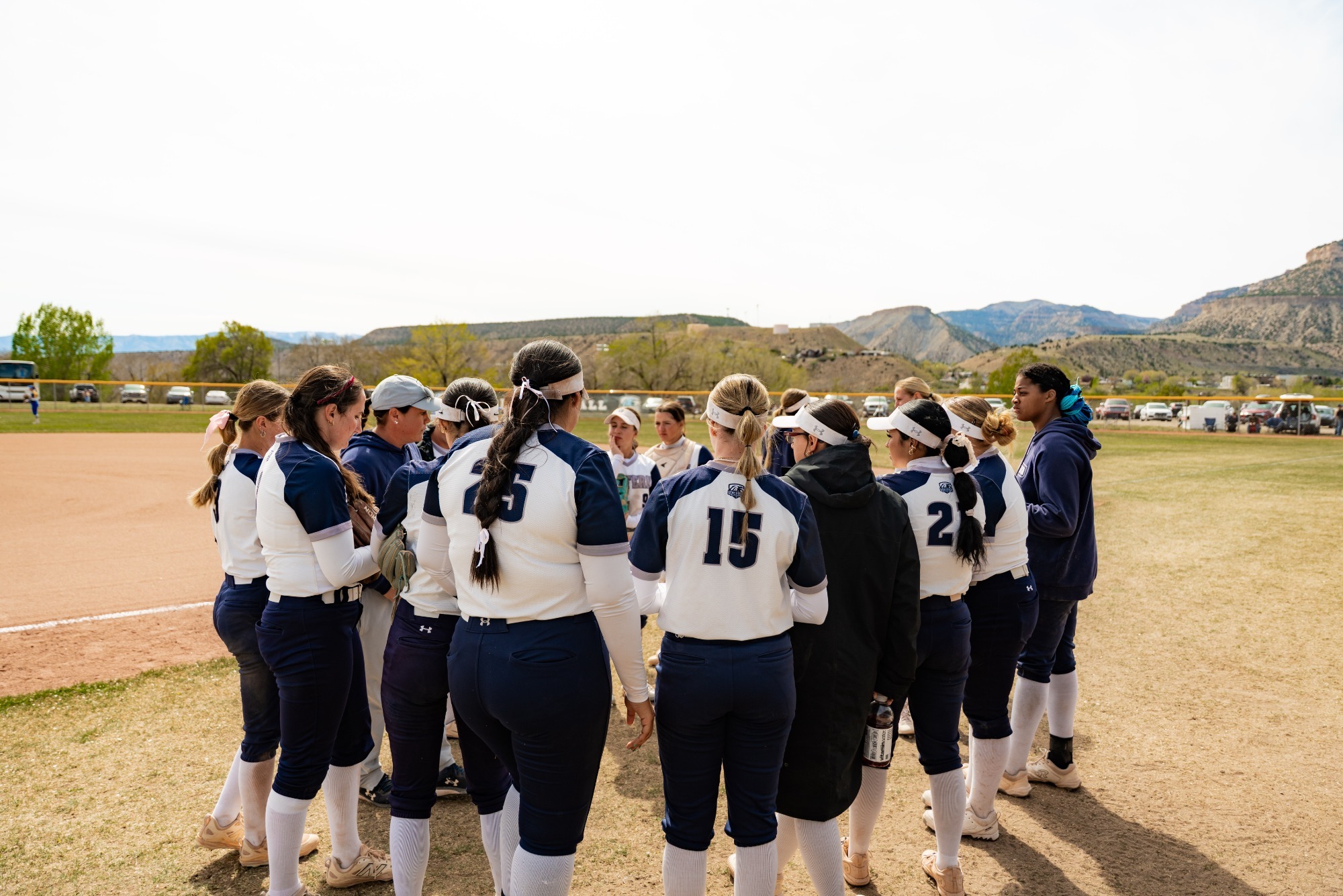 A large group of Utah State Eastern softball players, wearing white and navy blue uniforms, gather in a huddle on a sunny day. They are surrounded by coaches and staff on a dirt and grass field. In the background, the outfield fence, several parked cars, and large desert hills are visible under a bright sky.