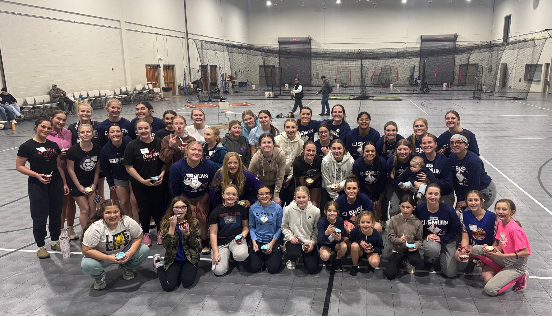 A wide group shot of approximately 40 people, primarily young female athletes and coaches, gathered on a grey indoor sports court. They are smiling for the camera, and several individuals in the front and middle rows are holding small blue and white cups. In the background, there is a large black batting cage net and gym equipment. The lighting is bright from overhead stadium-style lights.