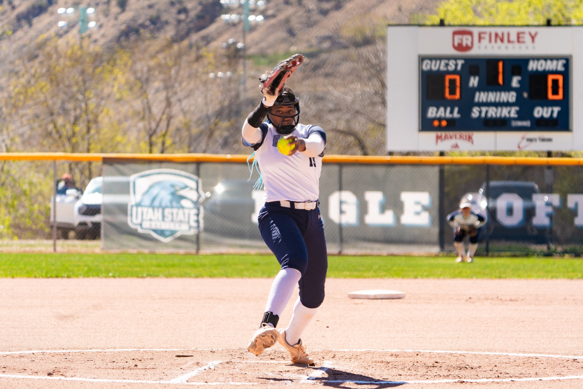Action shot of a softball pitcher delivering a pitch. She wears a white jersey, dark blue pants, and a protective face mask. In the background, an electronic scoreboard reads 
