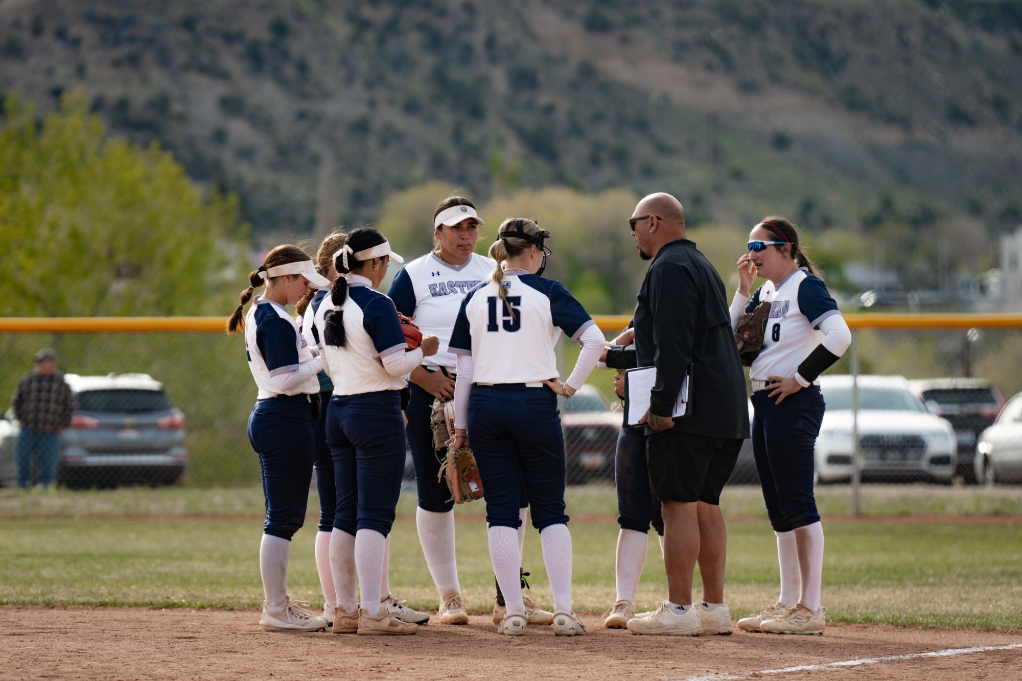 Six members of the Utah State Eastern softball team, wearing white and navy blue uniforms, gather in a huddle on the dirt infield. They are listening to a coach in a black shirt and shorts who is holding a clipboard. The background shows an outfield fence, parked cars, and a mountain under a clear sky.