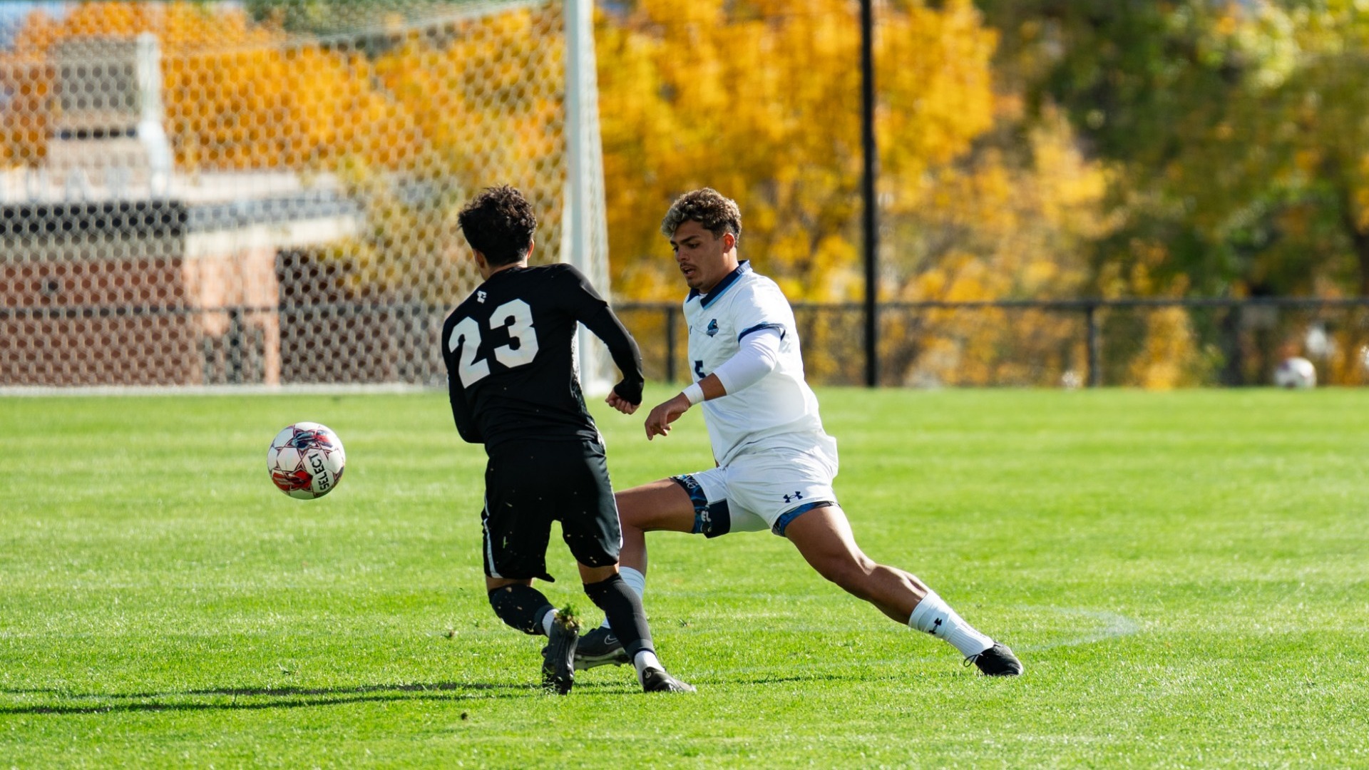 An outdoor action photo of a Utah State Eastern male soccer player in a white uniform extending his leg to control the ball while fending off an opposing defender wearing number 23 in black. A soccer goal and vibrant yellow autumn trees are visible in the background.