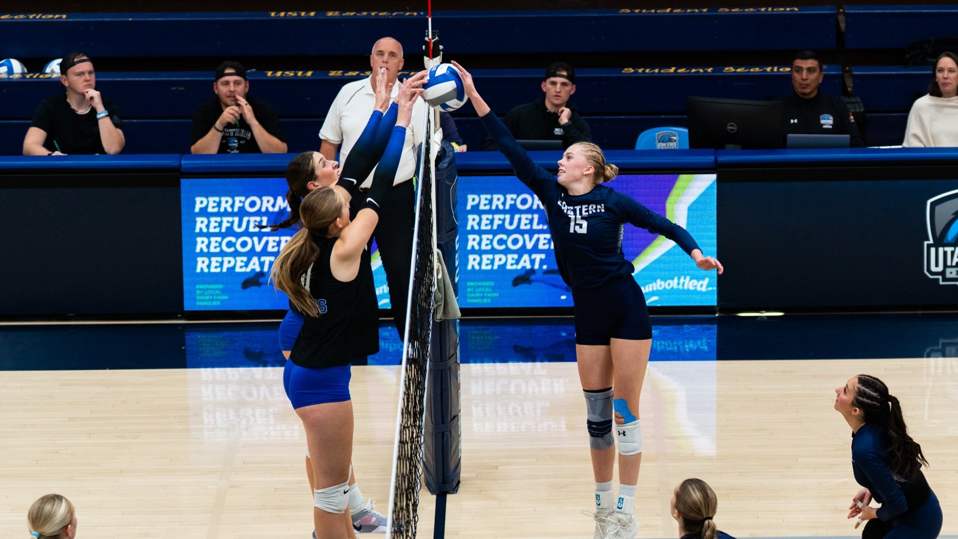 An indoor action photo of a Utah State Eastern volleyball player (wearing number 15 in a dark blue uniform) jumping high above the net to tip or block the ball. Two opposing players in black and blue uniforms jump on the other side of the net to contest the play, with spectators and a referee visible in the background bleachers.