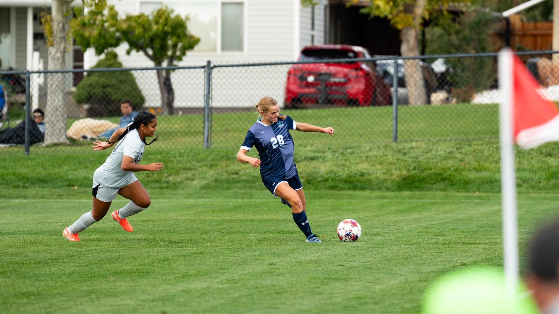 An action photo of a Utah State Eastern female soccer player (wearing number 28 in a navy blue jersey) dribbling a soccer ball along the sideline near a red corner flag, with an opposing player in a light grey uniform chasing close behind. A chain-link fence, parked cars, and trees are in the background.