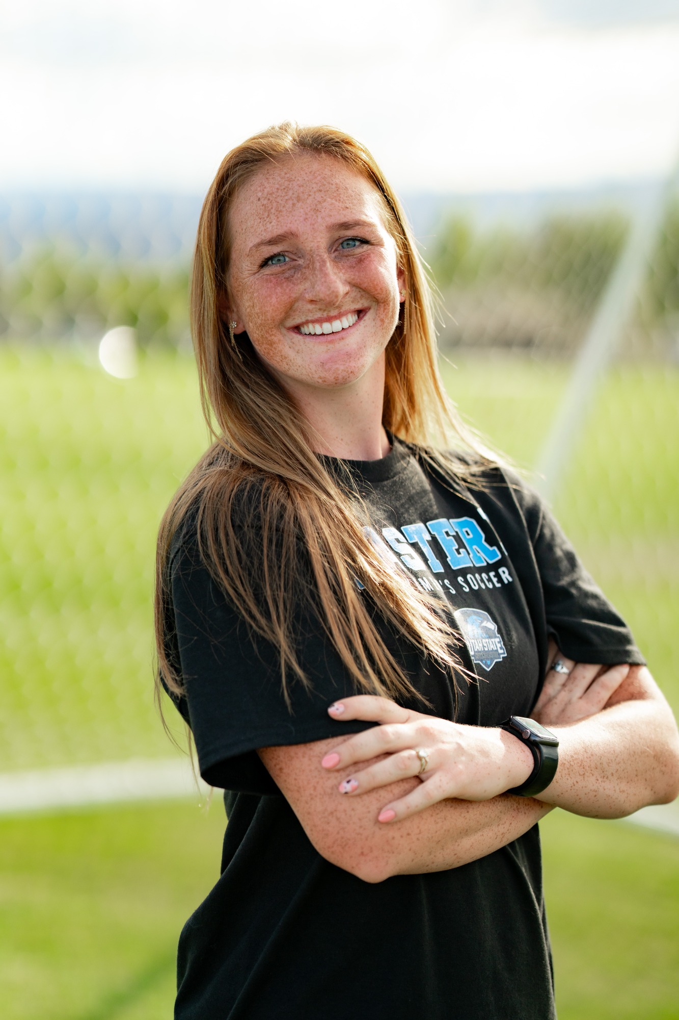 A smiling woman stands on a soccer field with her arms crossed, wearing a black Utah State Eastern soccer shirt