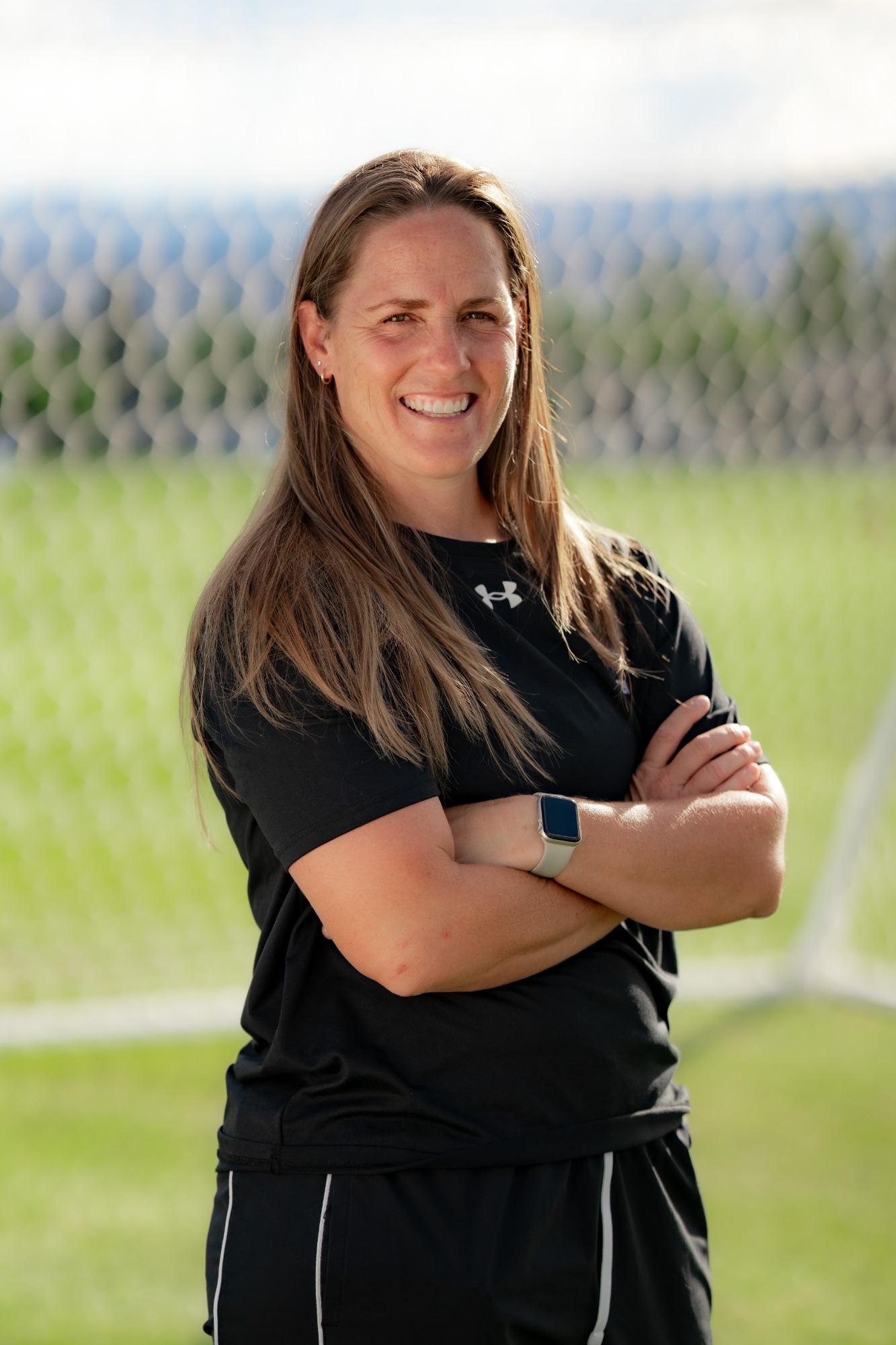 A smiling woman stands on a soccer field with her arms crossed, wearing a black athletic shirt and shorts