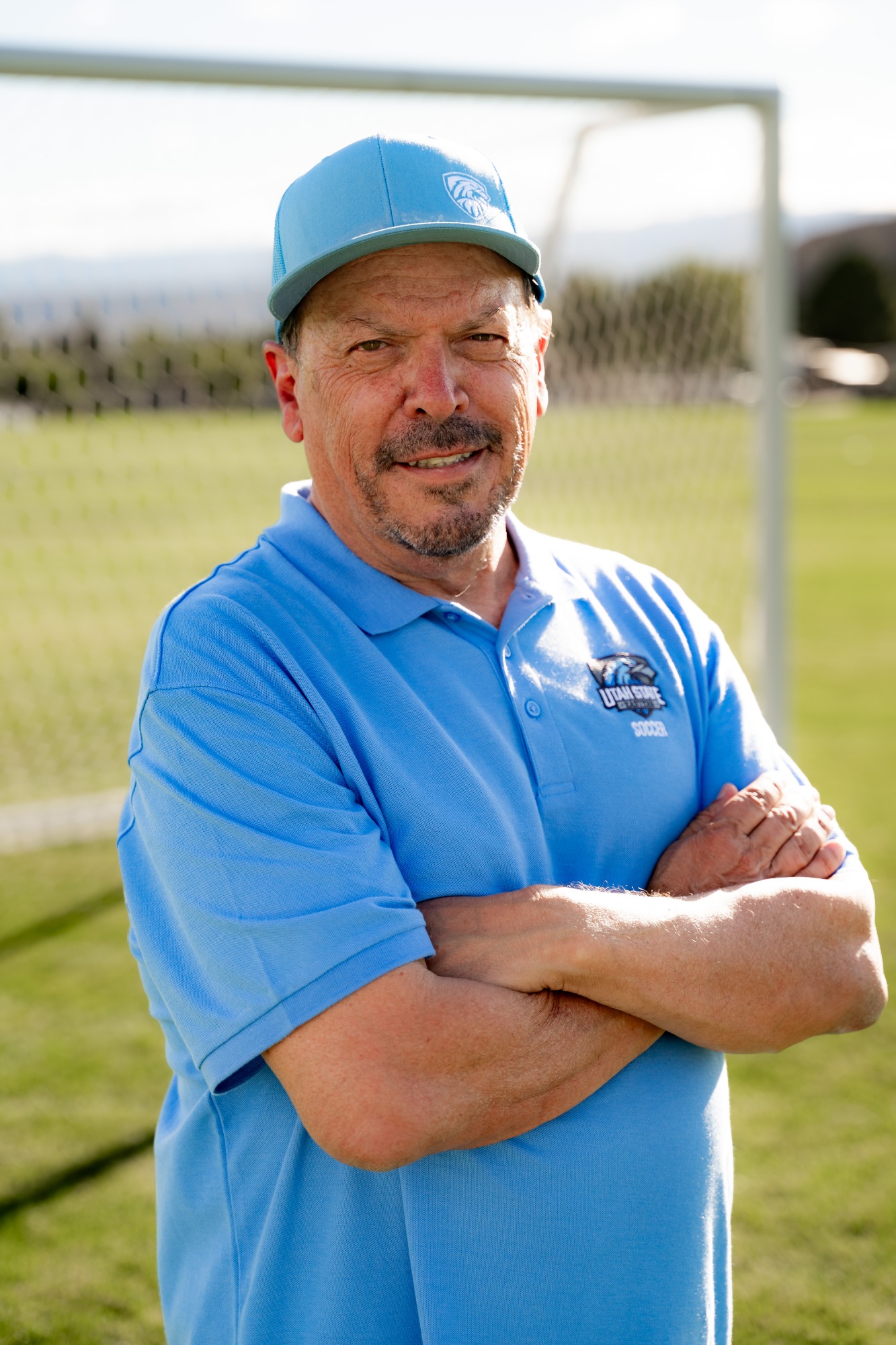 Utah State Eastern men’s soccer coach wearing a light blue Utah State Eastern polo and cap, standing with his arms crossed on the soccer field.
