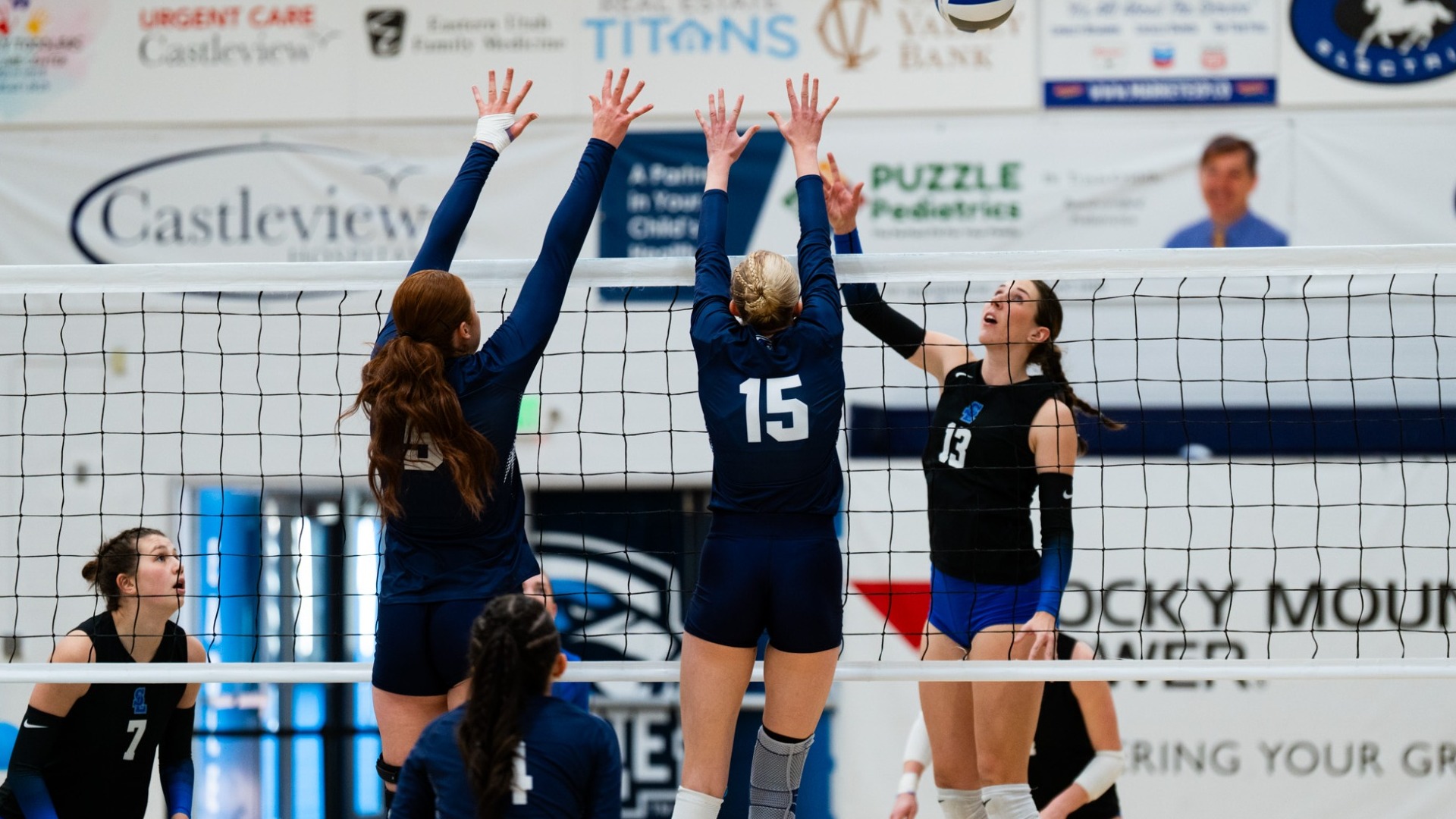 An indoor volleyball action shot captured at net level during a match. Two Utah State Eastern defenders wearing dark navy uniforms (including number 15) jump together with their arms fully extended above the net to form a wall against an opponent's attack. Across the net, an opposing hitter wearing a dark sleeveless jersey (number 13) leaps to strike the volleyball suspended in mid-air above her head. Additional teammates track the play from the court floor in the background.