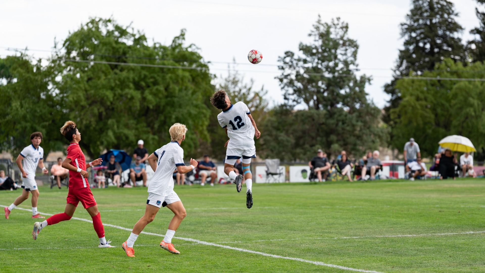 An outdoor sports photograph capturing a Utah State Eastern men's soccer player (number 12) jumping high off the grass pitch to head a soccer ball mid-air. Teammates in white uniforms and an opposing player in a red kit look up at the aerial play, with sideline spectators, umbrellas, and trees lining the background under an overcast sky.