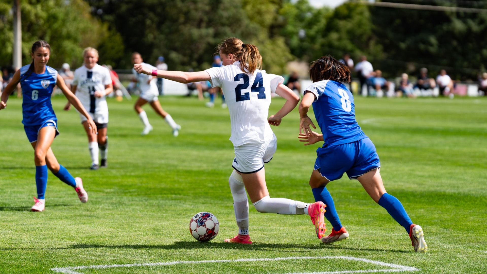 An outdoor action shot from a women's soccer match on a sunny day. A Utah State Eastern player in a white jersey (number 24) controls a soccer ball while driving past an opposing defender in a royal blue jersey (number 8). Additional players from both teams track the play across the green field, with lush green trees and spectators framing the background.