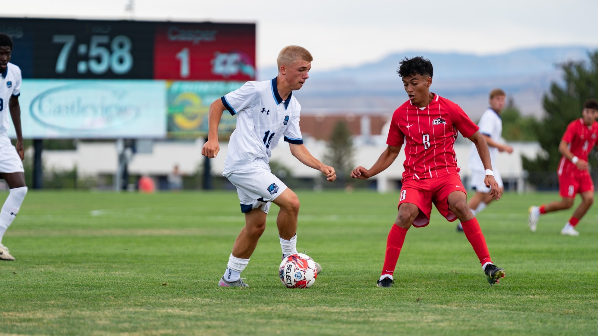 An outdoor match photo showing a blonde Utah State Eastern male soccer player in a white uniform with dark trim controlling the ball mid-dribble. A defender in a red uniform with pinstripes (number 8) stays low in a defensive stance facing him. An electronic scoreboard displaying 