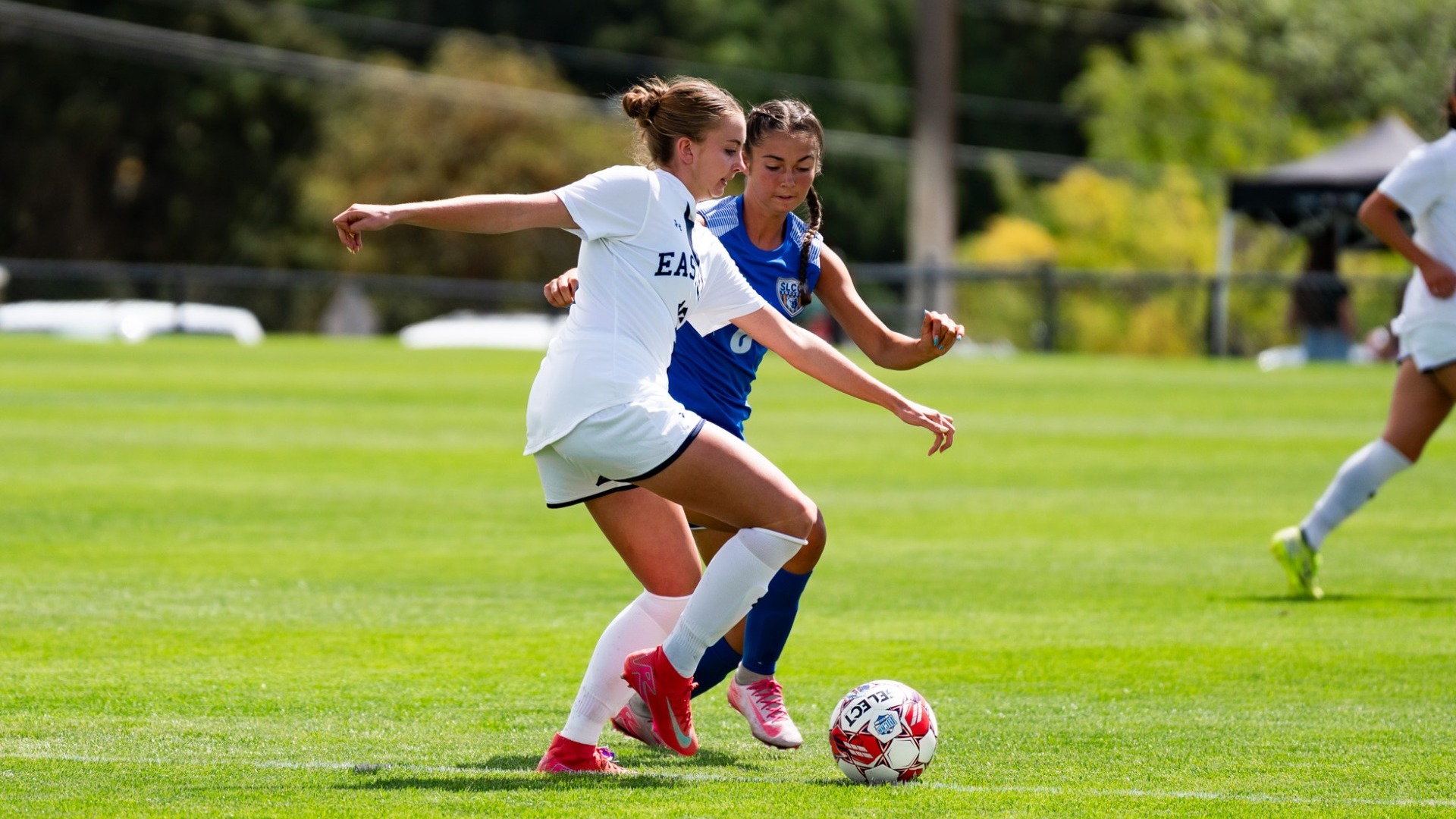 An outdoor action photograph from a women's soccer match on a sunny day. A Utah State Eastern player in a white jersey (number 16) and matching white shorts extends her arm to shield a soccer ball from a closely contesting defender wearing a royal blue uniform. Another teammate in white runs parallel down the field in the background behind a line of mature green trees.
