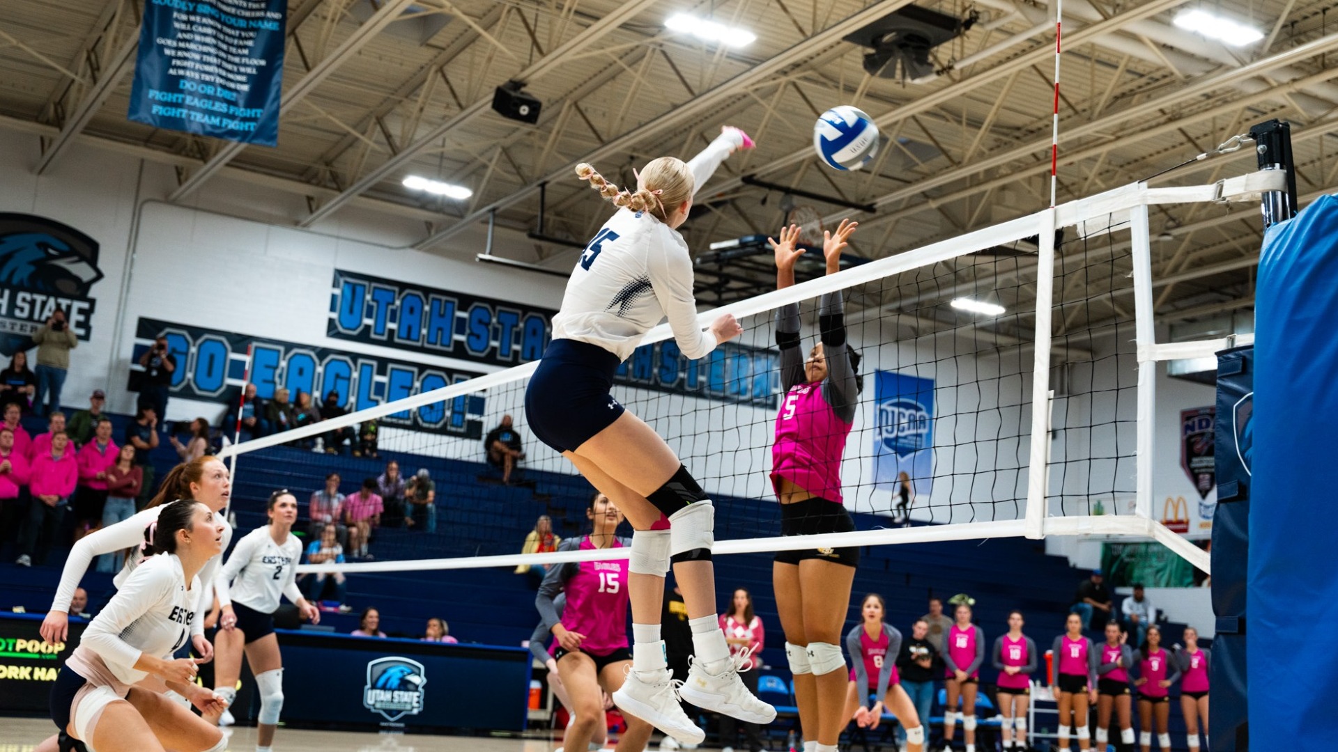 An indoor action photograph from a volleyball match inside the Utah State Eastern gymnasium. A Utah State Eastern player with blonde braided hair (number 15), wearing a white long-sleeve jersey and dark navy shorts, jumps high into the air at the net to hit a blue and white volleyball. Across the net, an opposing blocker in a bright pink jersey reaches up with both hands to attempt a block. Teammates watch the play from the court while spectators, banners, and blue bleachers fill the background.