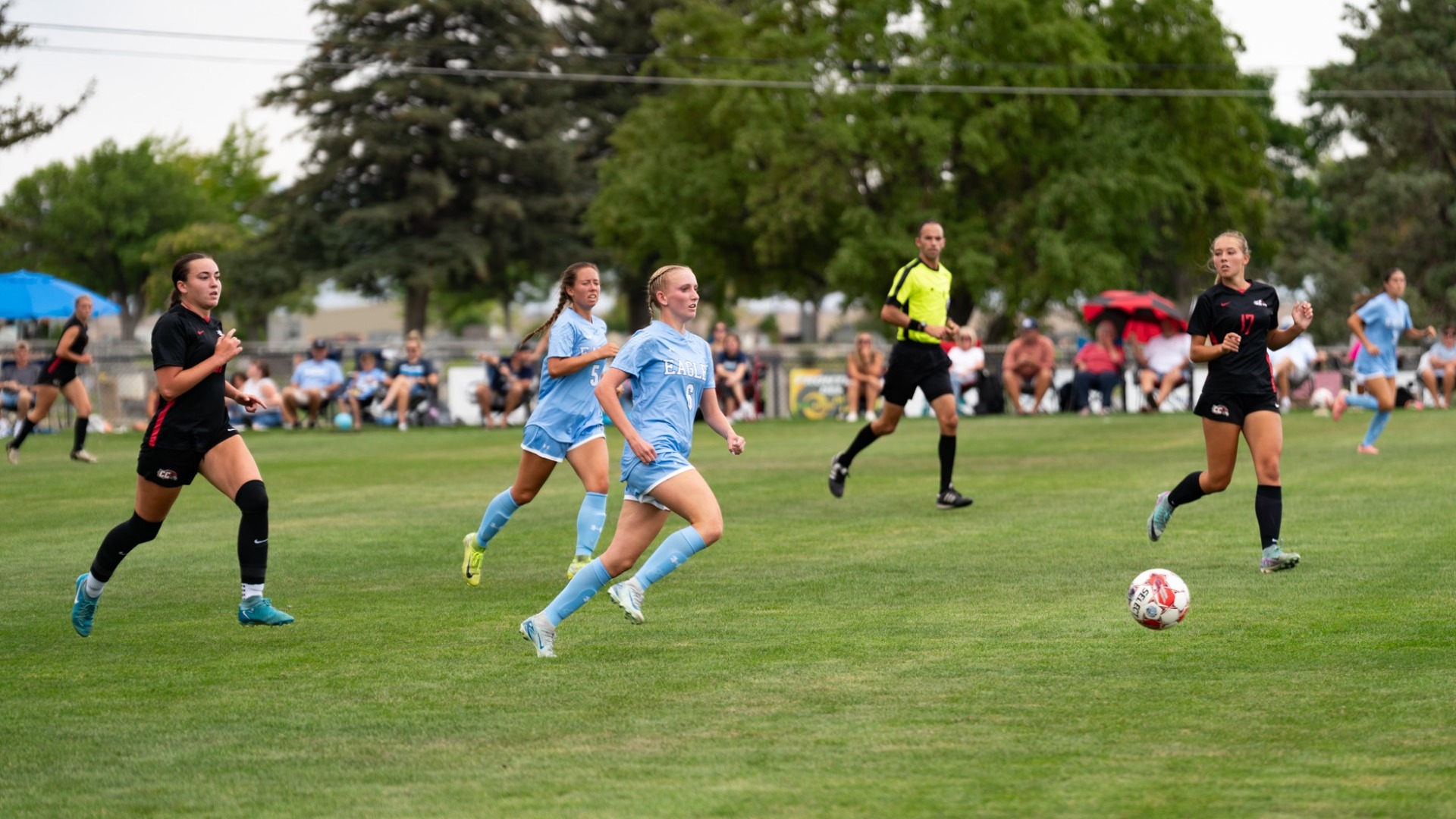 An action shot from a women's soccer match on an outdoor grass field. A Utah State Eastern player (number 6) in a light blue uniform sprints toward a soccer ball rolling across the grass, flanked by a teammate (number 5) also in light blue. Two opposing players in black uniforms move into defensive position nearby, with a referee in a neon yellow shirt trailing behind the play. Trees and spectators lined along the fence frame the background.