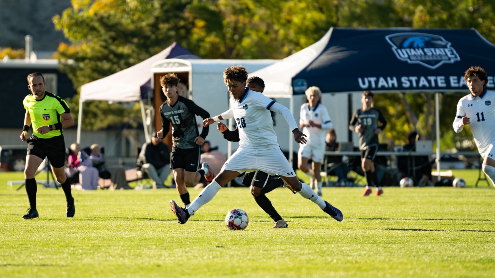 An outdoor action photograph from a men's soccer match on a bright afternoon. A Utah State Eastern player in a white uniform with dark trim (number 28) lunges low to control the ball between his feet while fending off an opposing defender in a black kit. In the background, a referee in bright yellow tracks the play, white team canopy tents sit along the sideline, and additional players watch from the pitch and bench.