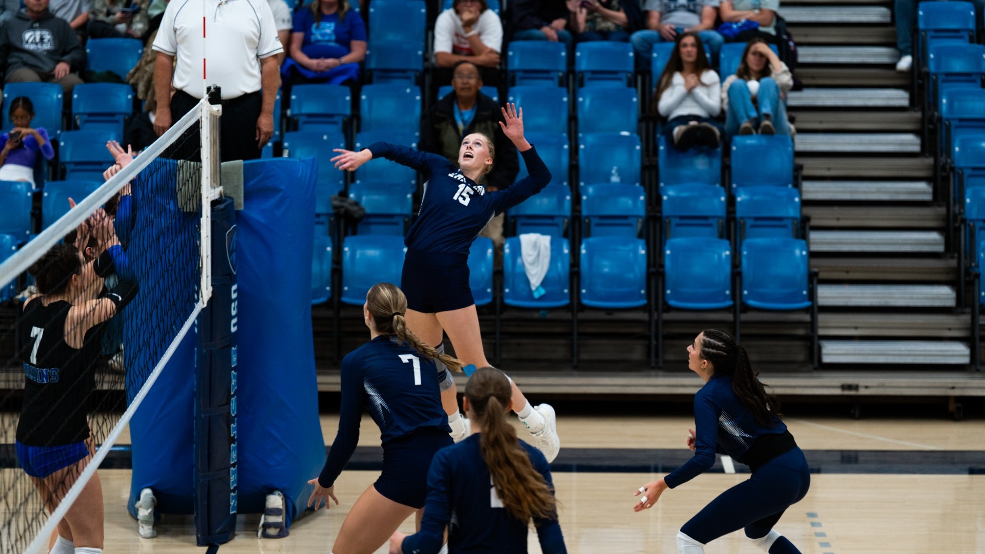 An indoor action photo taken from the side of a volleyball court. A Utah State Eastern player in a dark navy long-sleeve uniform (number 15) leaps into the air near the net with her arm cocked back to hit the ball. Teammates in matching dark navy uniforms get into position below her, while an opposing player (number 7) watches from across the net. A referee stands on the elevated platform on the left, and spectators fill the blue arena seats behind.