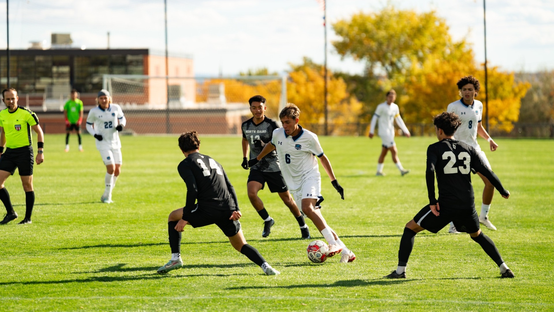 An outdoor action shot from a men's soccer match played on a grass field backed by bright yellow autumn trees. A Utah State Eastern player wearing a white jersey (number 6) dribbles the ball near mid-field while surrounded by two opposing players in black kits (numbers 14 and 23). A referee in a high-visibility yellow shirt tracks the action from the left, while other players position themselves nearby under clear skies.