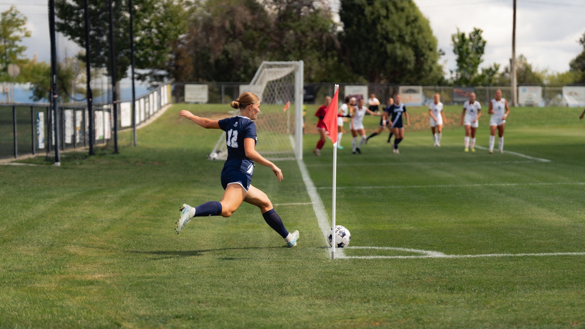 An action photograph of a Utah State Eastern women's soccer player (number 12) in a dark navy uniform taking a corner kick. She is winding up her right leg near the red corner flag, with the soccer ball placed on the corner arc line. Other players from both teams wait in the penalty box in the background, set against an outdoor pitch with trees and a goal frame behind the goal line.