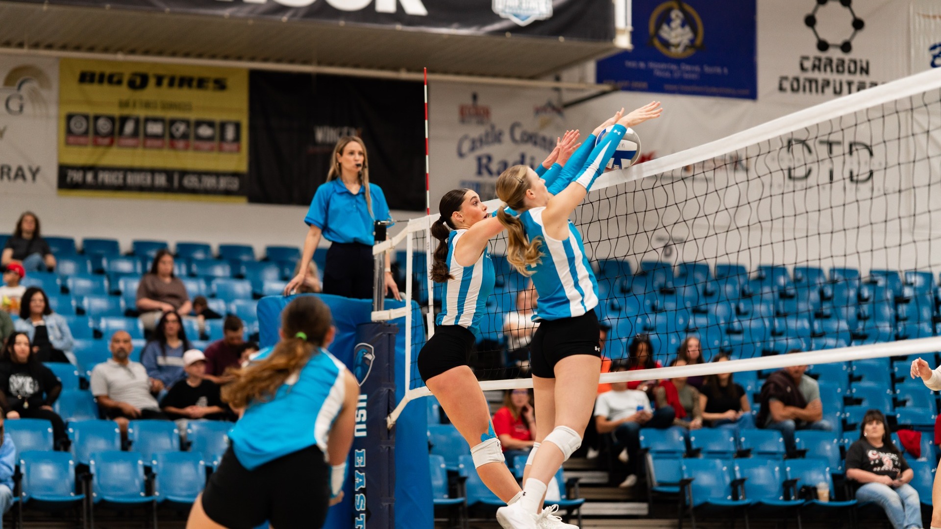 An indoor action photo showing two Utah State Eastern volleyball players in light blue and white jerseys jumping simultaneously at the net, extending their arms to block an opponent's attack. A teammate in light blue watches the play from the foreground, while the referee sits in the background platform chair. Blue stadium seating and sponsor banners are visible behind the court.