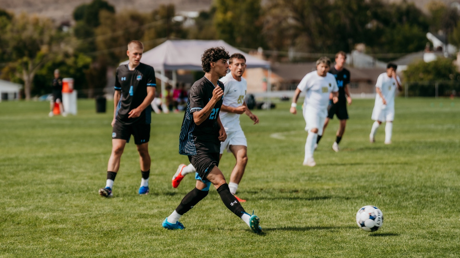 An outdoor action shot of a Utah State Eastern men's soccer player (number 21) in a dark blue kit dribbling the ball down the field during a match against TMCC. Teammates and opposing players in white jerseys track the play in the background across the green pitch, with foothills visible in the distance.