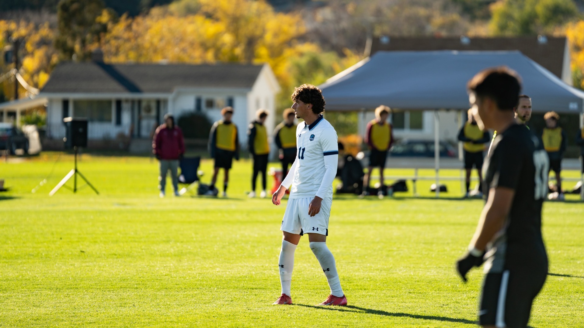 An outdoor action photograph from a men's soccer match on a sunny field with autumn foliage and residential houses in the background. A Utah State Eastern player with curly dark hair wearing a white long-sleeve jersey with dark trim (number 11) and matching white shorts stands in profile on the bright green grass. An opponent in a dark jersey stands in the foreground, while additional players, team personnel, and a sideline canopy tent line the background.