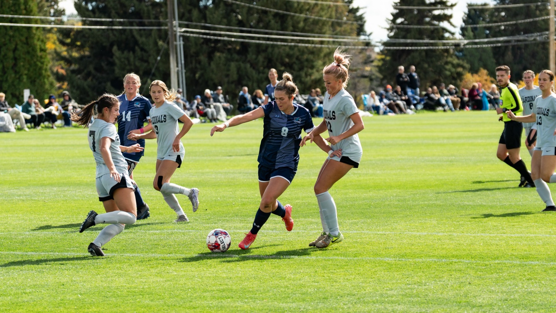 An outdoor action photo from a women's soccer match on a sunny day. A Utah State Eastern player in a dark navy uniform (number 8) controls the ball mid-field, maneuvering past two opposing defenders in light gray jerseys (numbers 7 and 22). Additional teammates and defenders surround the play, while spectators and a match referee line the grassy background near the tree-lined boundary.