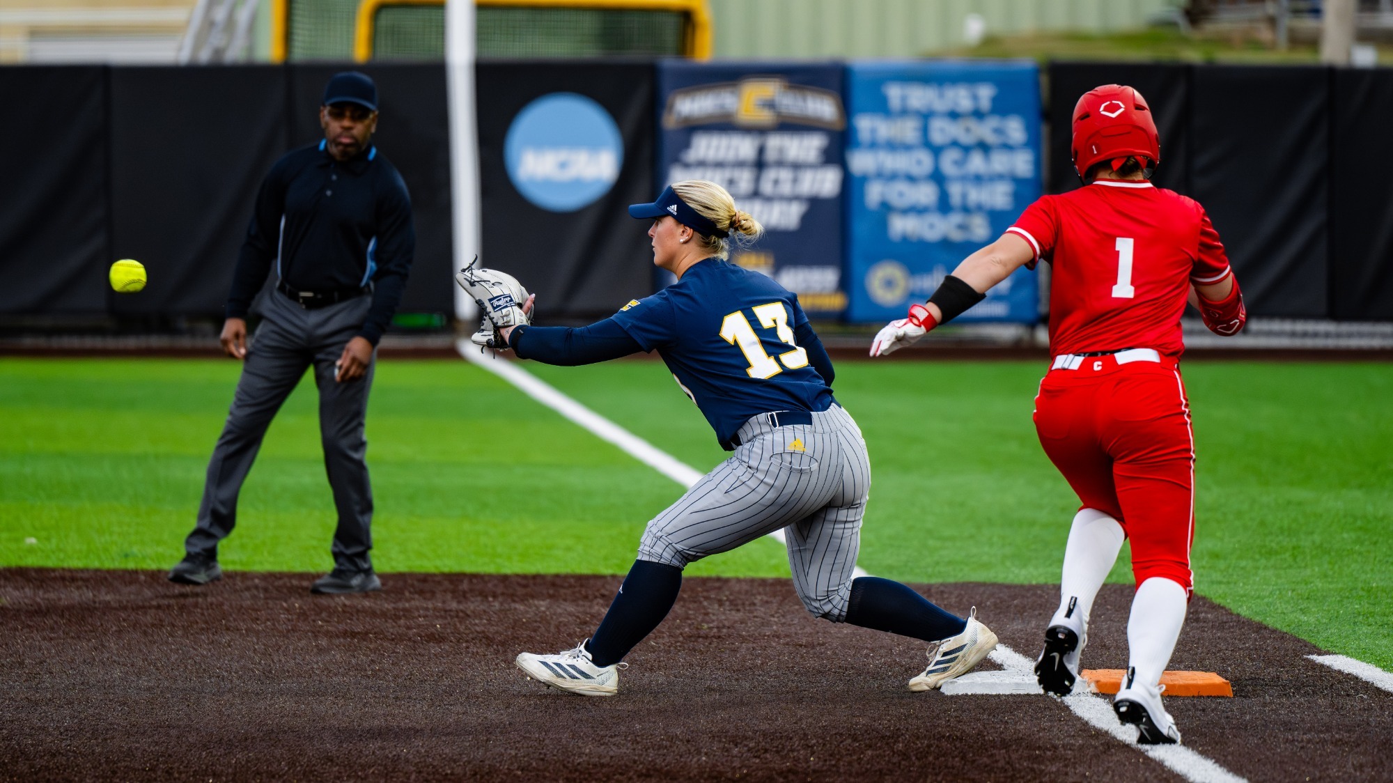 A softball player catches the throw to make an out at first base with another player running to first.