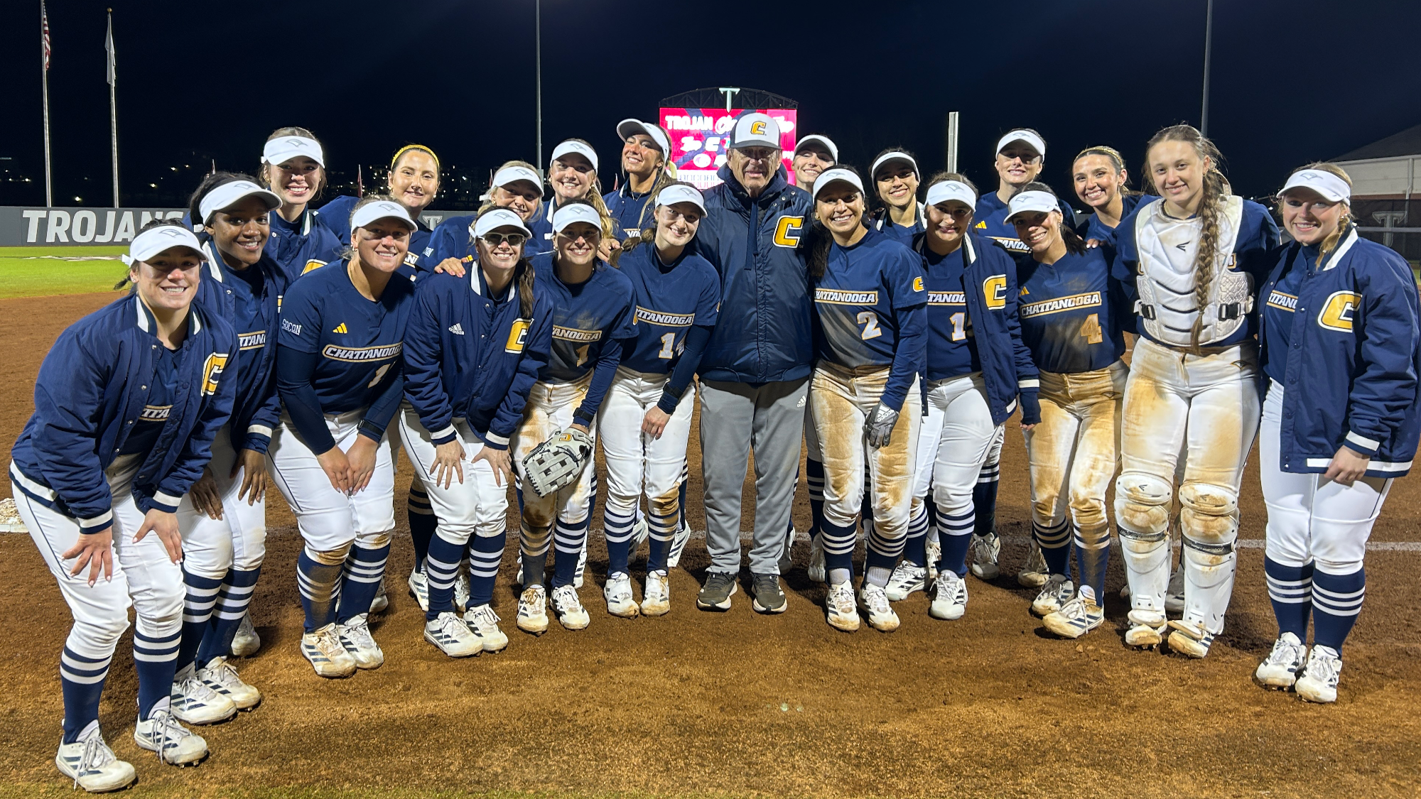Softball team poses with head coach on the softball field to celebrate his 800th career win