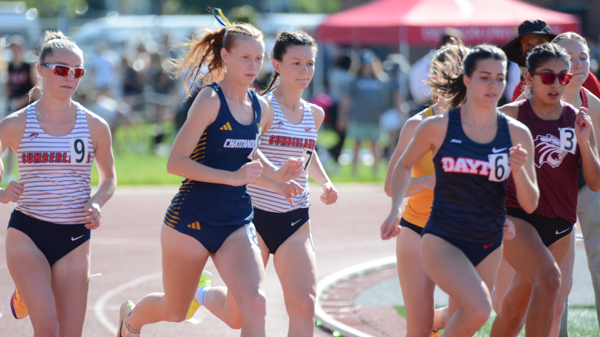 A Chattanooga runner in a navy and gold uniform races on an outdoor track alongside competitors from several teams during a mid-distance event, with spectators and tents visible in the background.