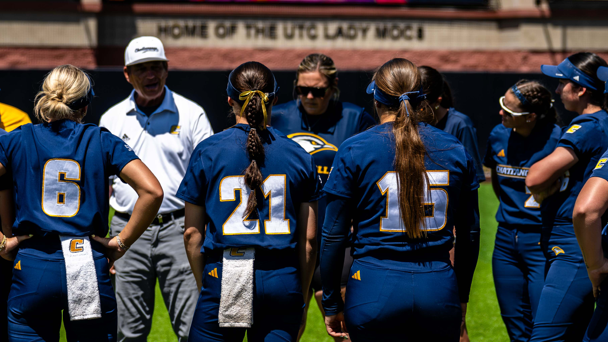 College softball players in navy blue uniforms stand in a huddle on a sunny field, listening to the male head coach before a game, with “Home of the UTC Lady Mocs” visible on the scoreboard behind them.
