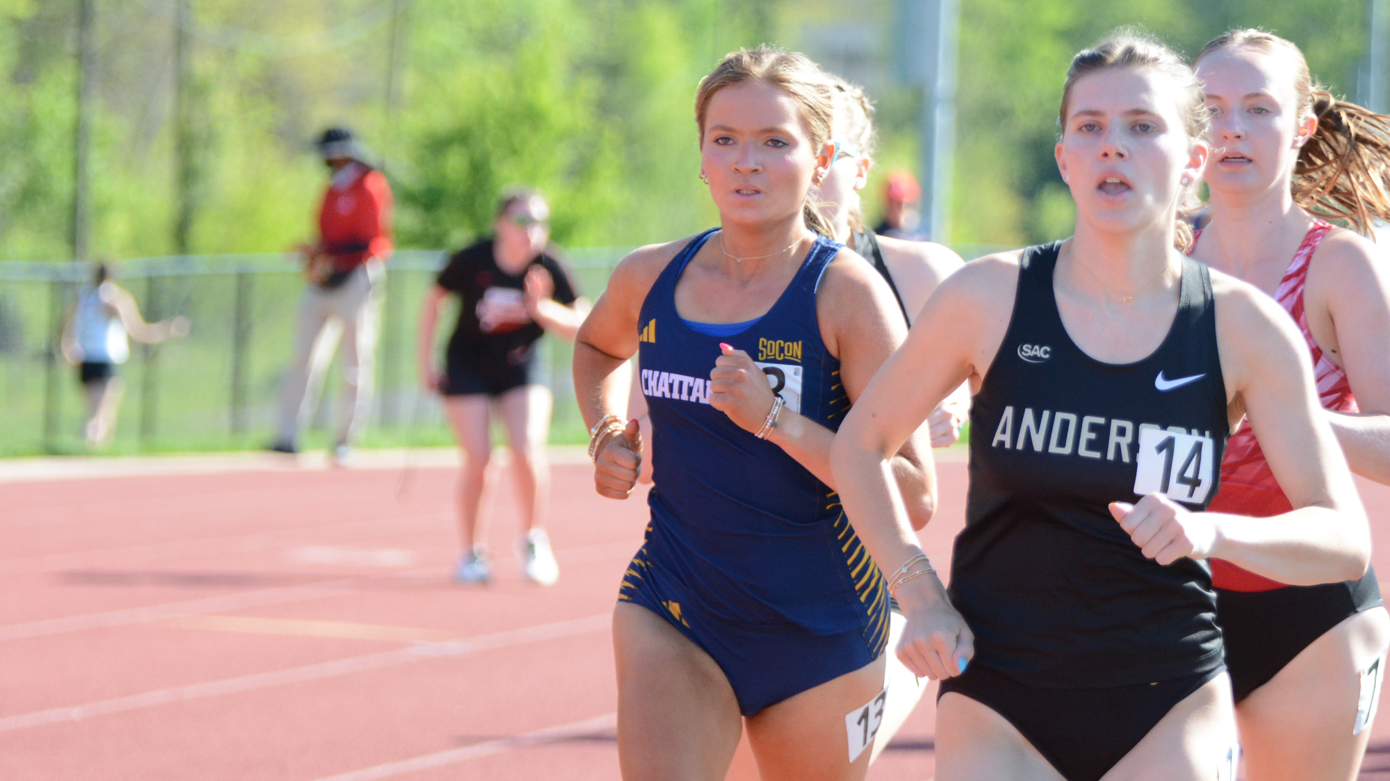 A Chattanooga Mocs track athlete in a navy uniform runs on an outdoor track during a race, alongside competitors from other teams in daylight conditions.