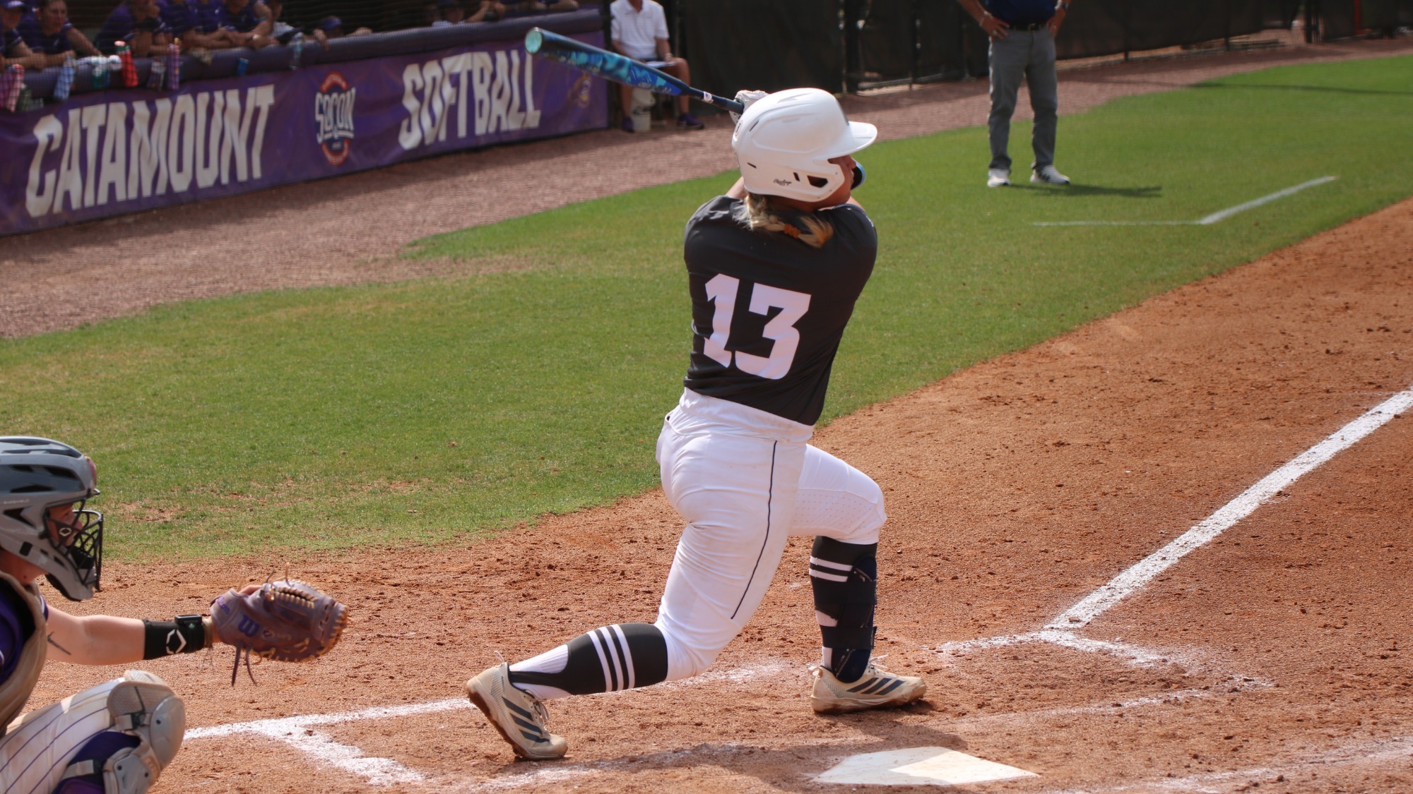 A Chattanooga Mocs softball player wearing a dark gray jersey with number 13 and a white helmet swings at a pitch at home plate while a catcher and umpire are positioned behind, with the Western Carolina dugout visible in the background.