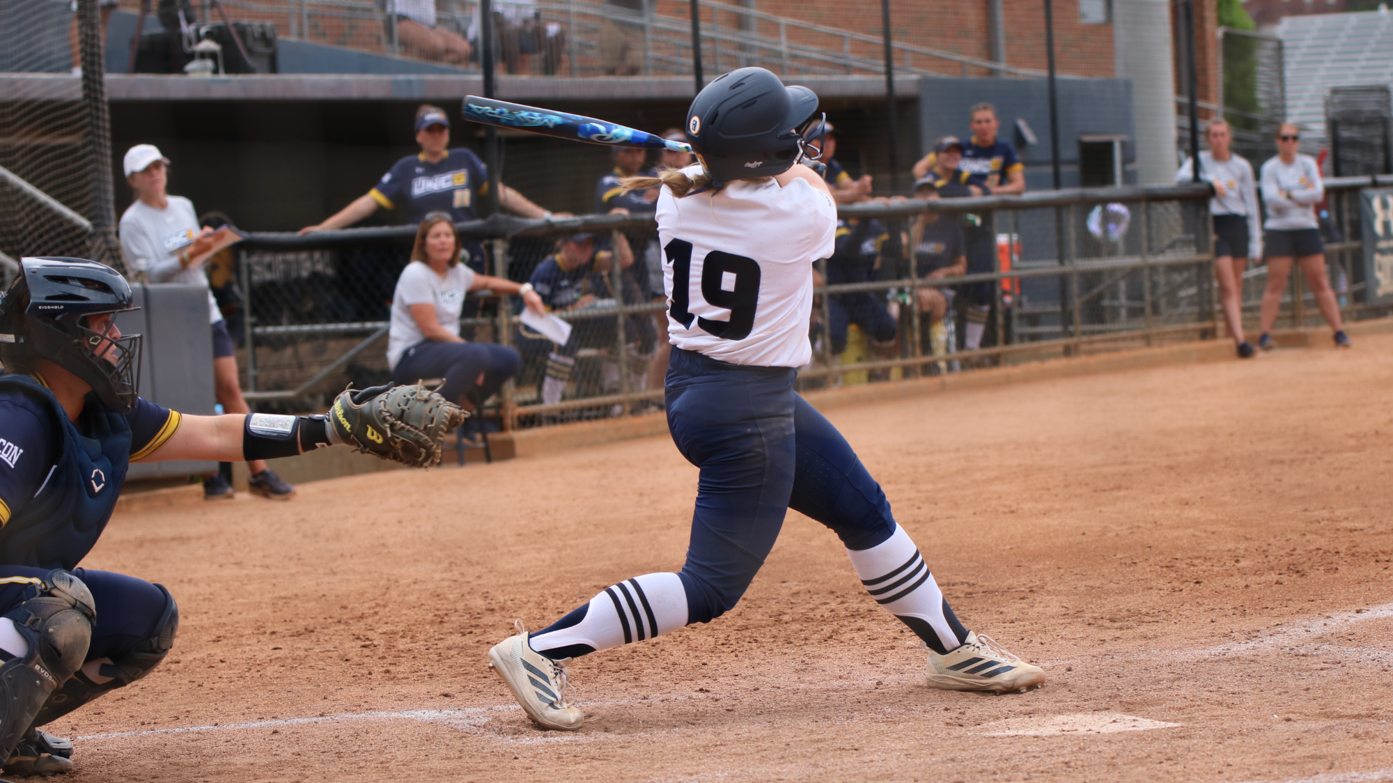 A softball player wearing a white jersey and blue pants is at the plate swinging at the ball for a hit