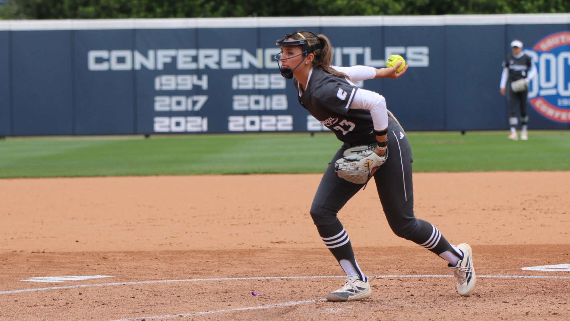 A softball pitcher in a dark uniform delivers a pitch from the infield during a game, holding a yellow softball mid-motion. Conference championship years are displayed on the outfield wall in the background, with another player visible near the outfield.