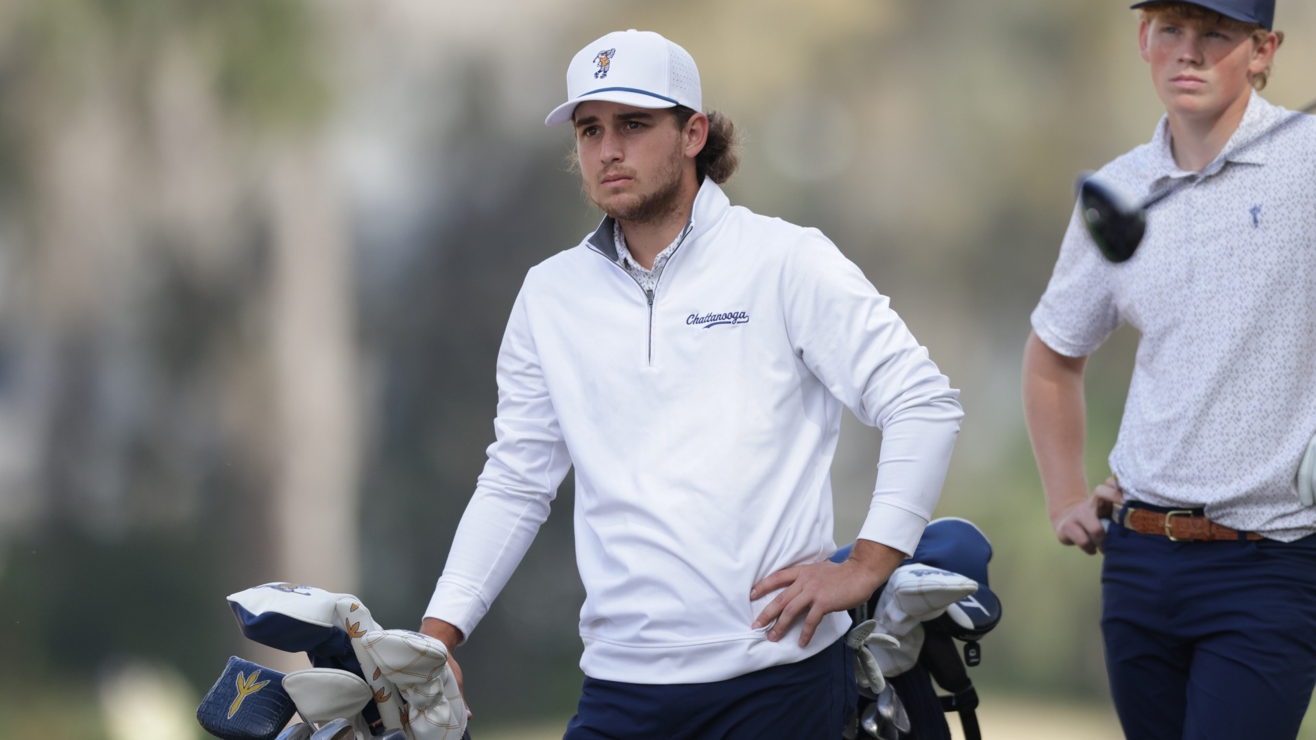 Ethan Whitaker looks down the fairway at his ball from the tee with a hand on his bag as he awaits playing partner's shot.