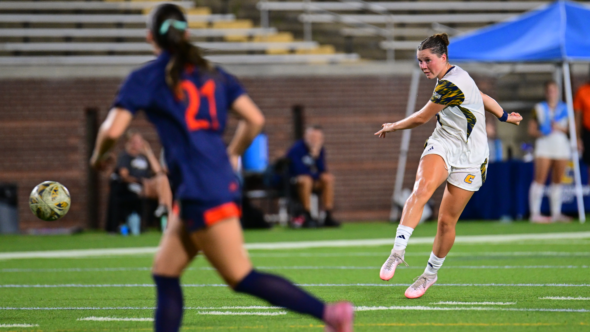 A soccer player in a white uniform kicks the ball downfield during a match while an opposing player in a navy uniform stands in the foreground.