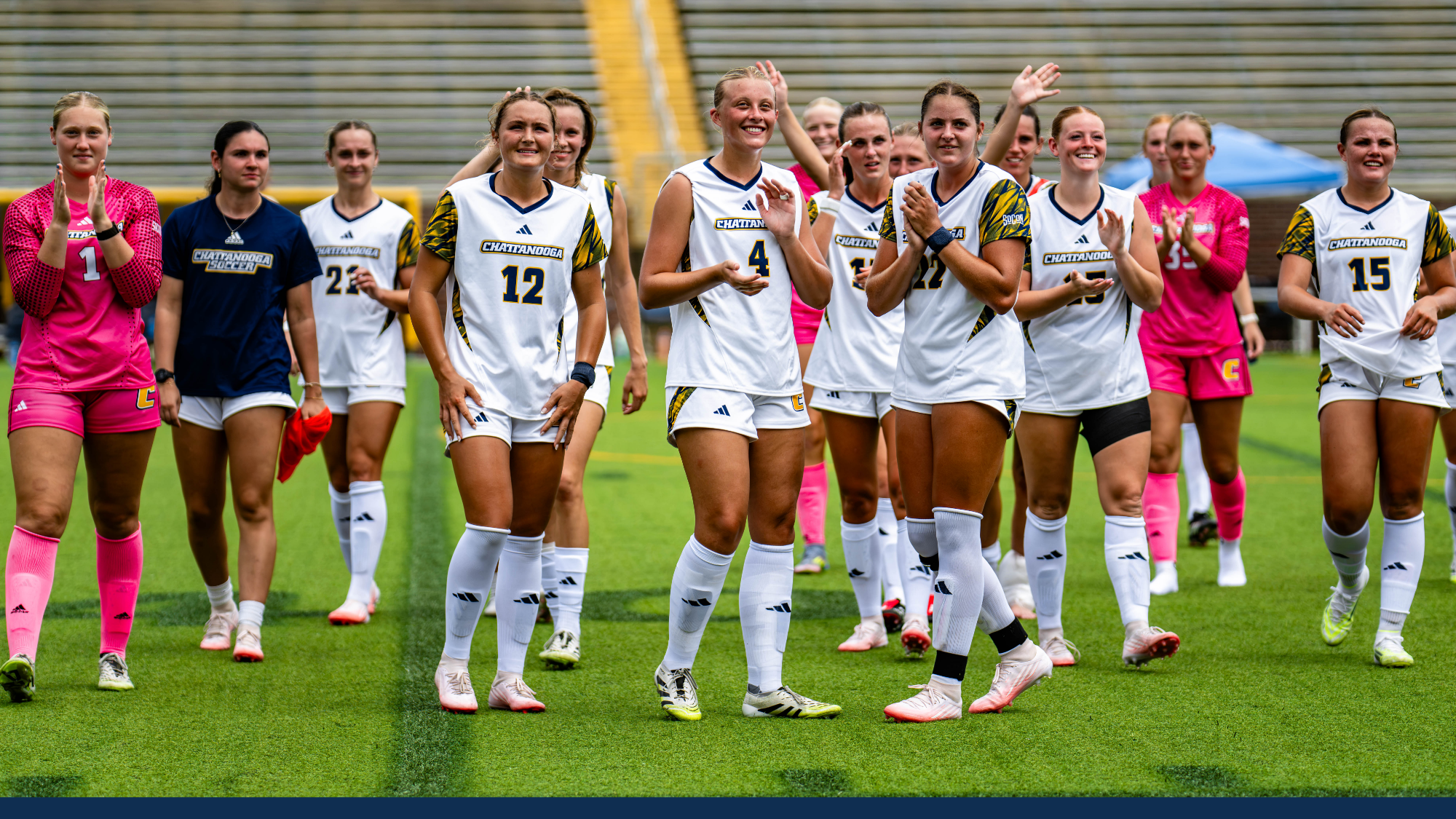 Several women's soccer players on the pitch are celebrating a win and waving at the crowd after the match.