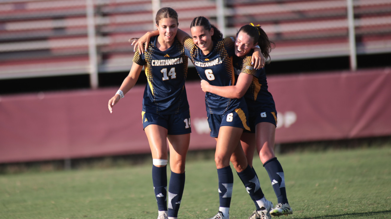 Three Chattanooga soccer players in navy and gold uniforms walk arm-in-arm on the field during a match. The player wearing No. 6 is between teammates wearing No. 14 and another teammate, with stadium bleachers visible in the background.