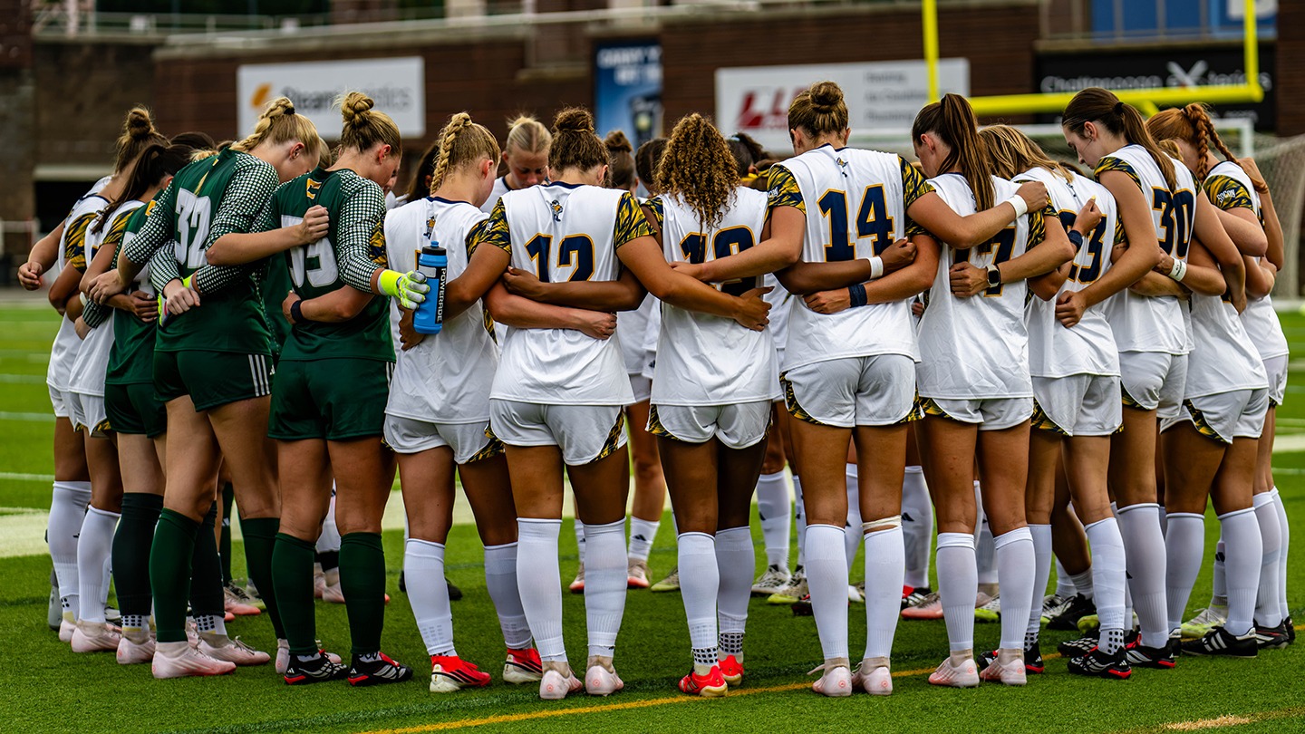 A soccer team in white and goalie green huddles together on the field before a game.