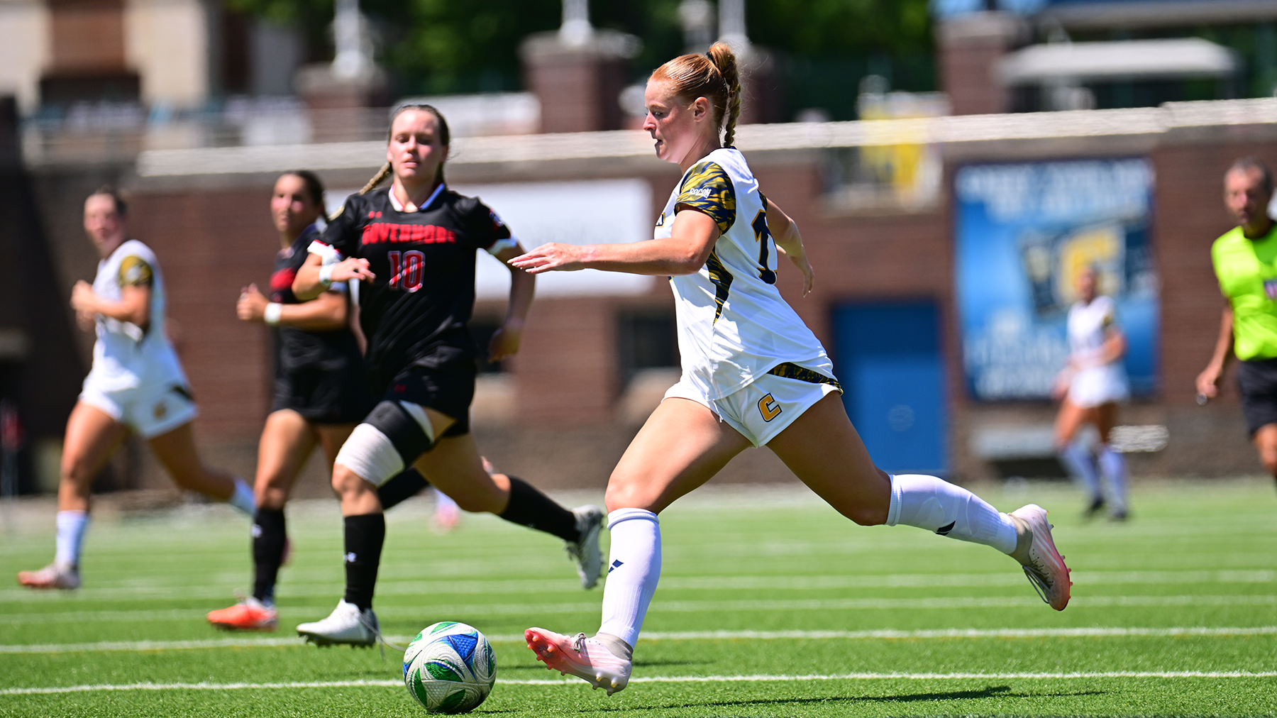 A women's soccer player in a white uniform dribbles the ball upfield during a match while two defenders in black uniforms pursue. The play takes place on a sunny afternoon with other players and an official visible in the background.