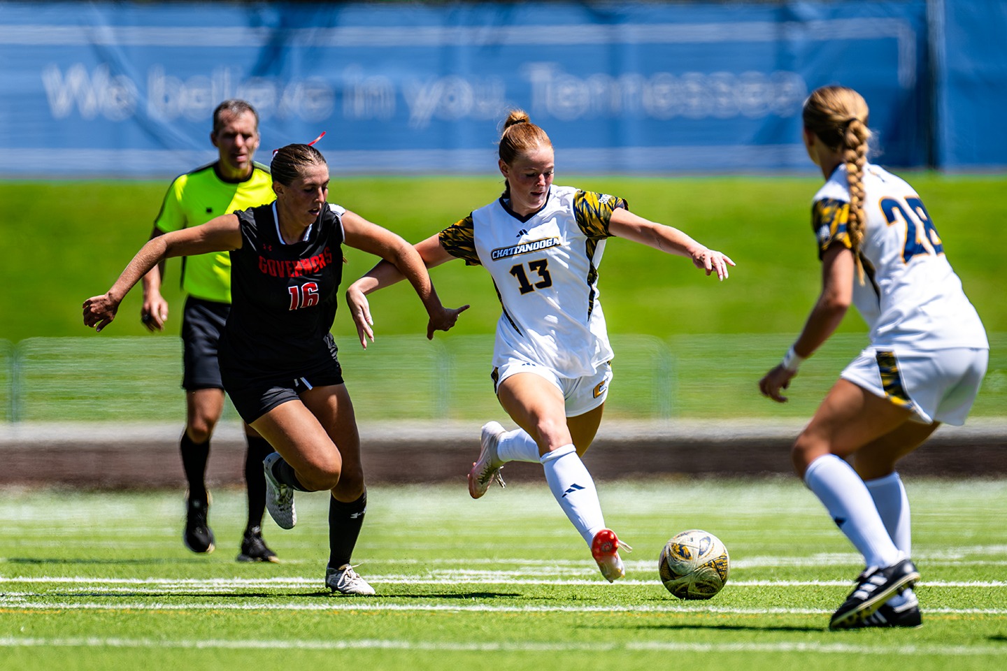 A soccer player wearing white is dribbling the ball while being pursued by a soccer player in black on the pitch.