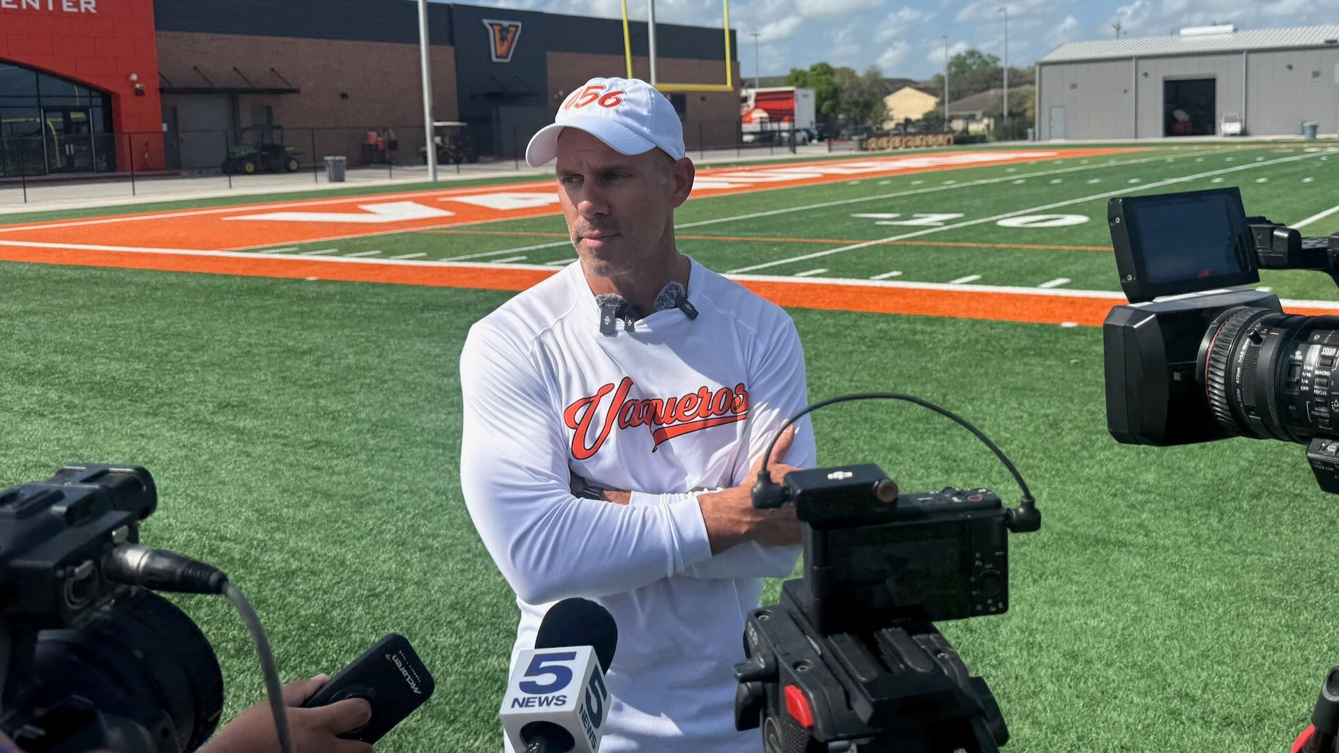 Image shows head utrgv football coach Travis Bush in a white shirt and white cap standing in front of cameras during a press conference on the turf.
