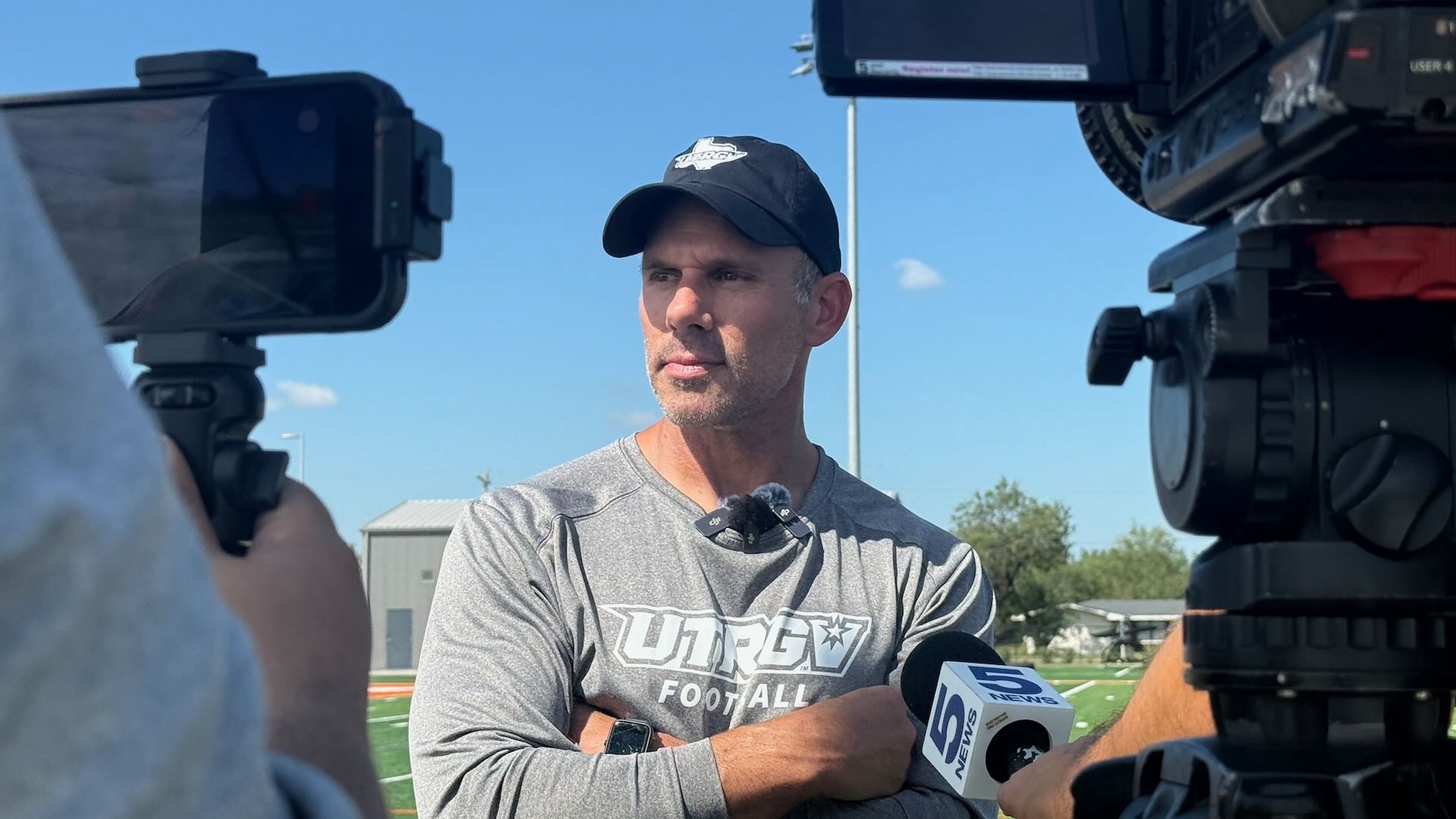 Image shows head coach Travis Bush in a black cap and gray shirt standing in front of cameras.
