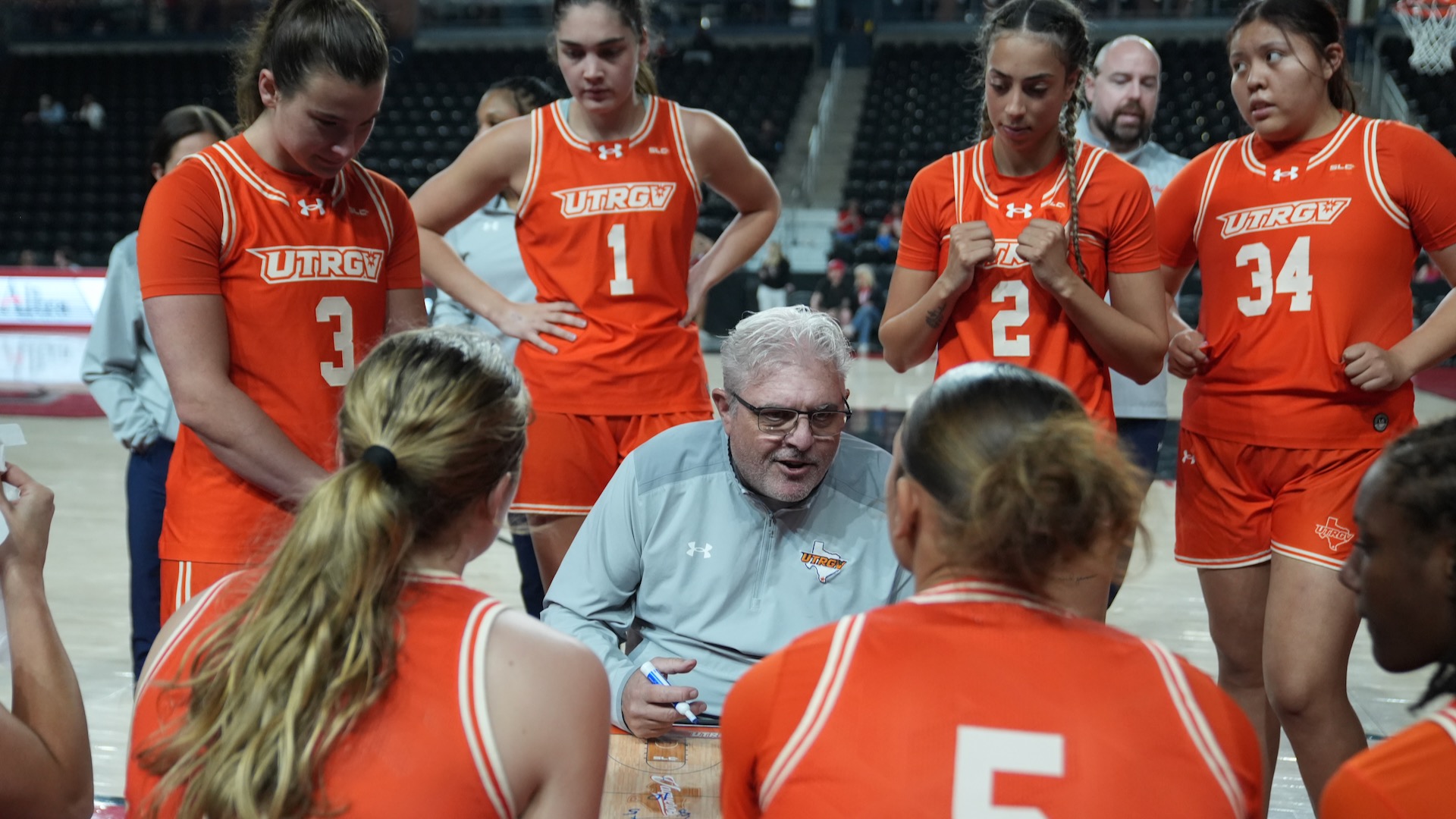 Head coach Lane Lord sits in the center of a team huddle with a dry erase board while the UTRGV women's basketball team sits on the bench or stands behind him listening to the play