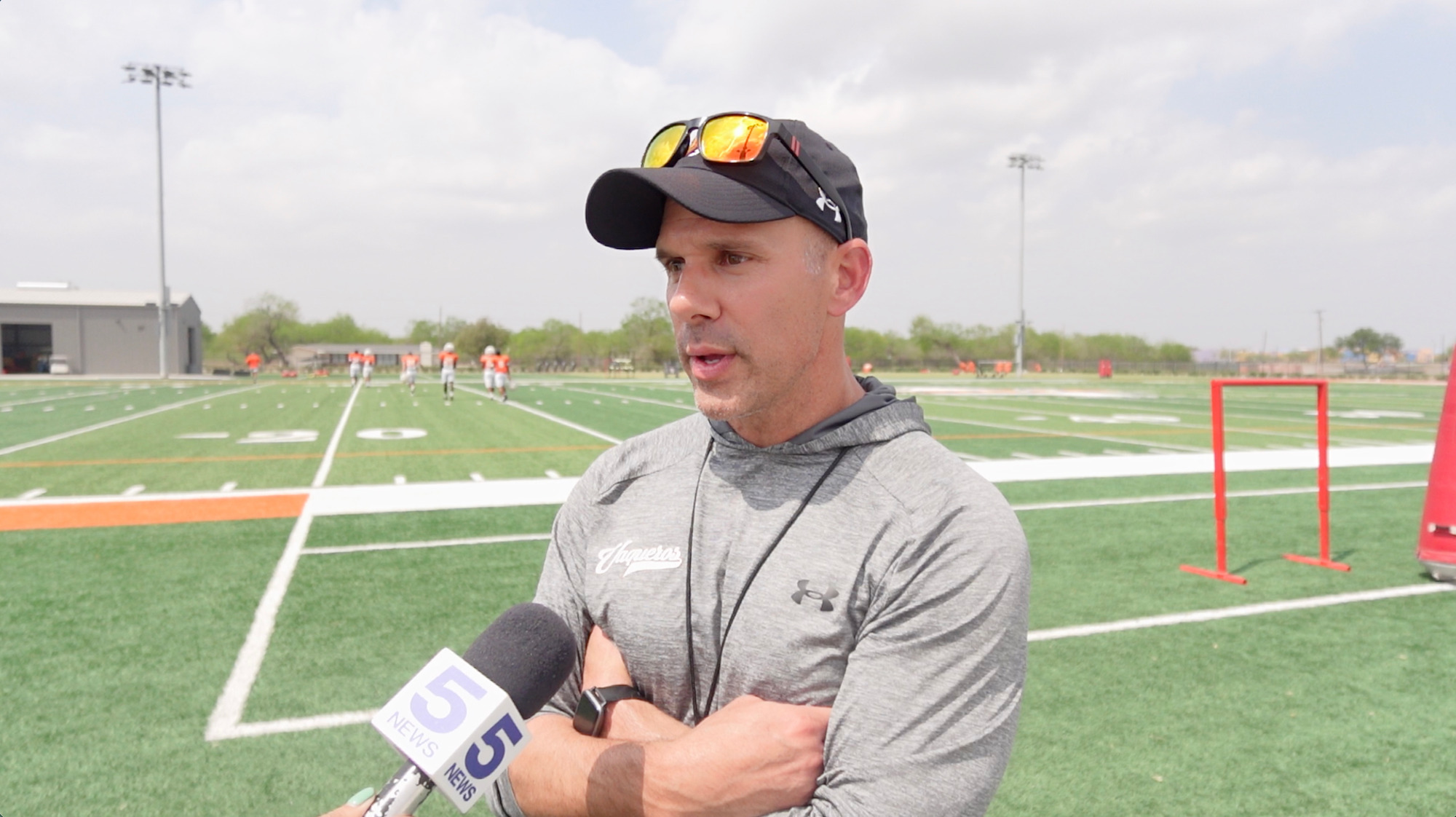 Coach Bush speaking with channel 5 with the turf practice field behind him 3-7-26