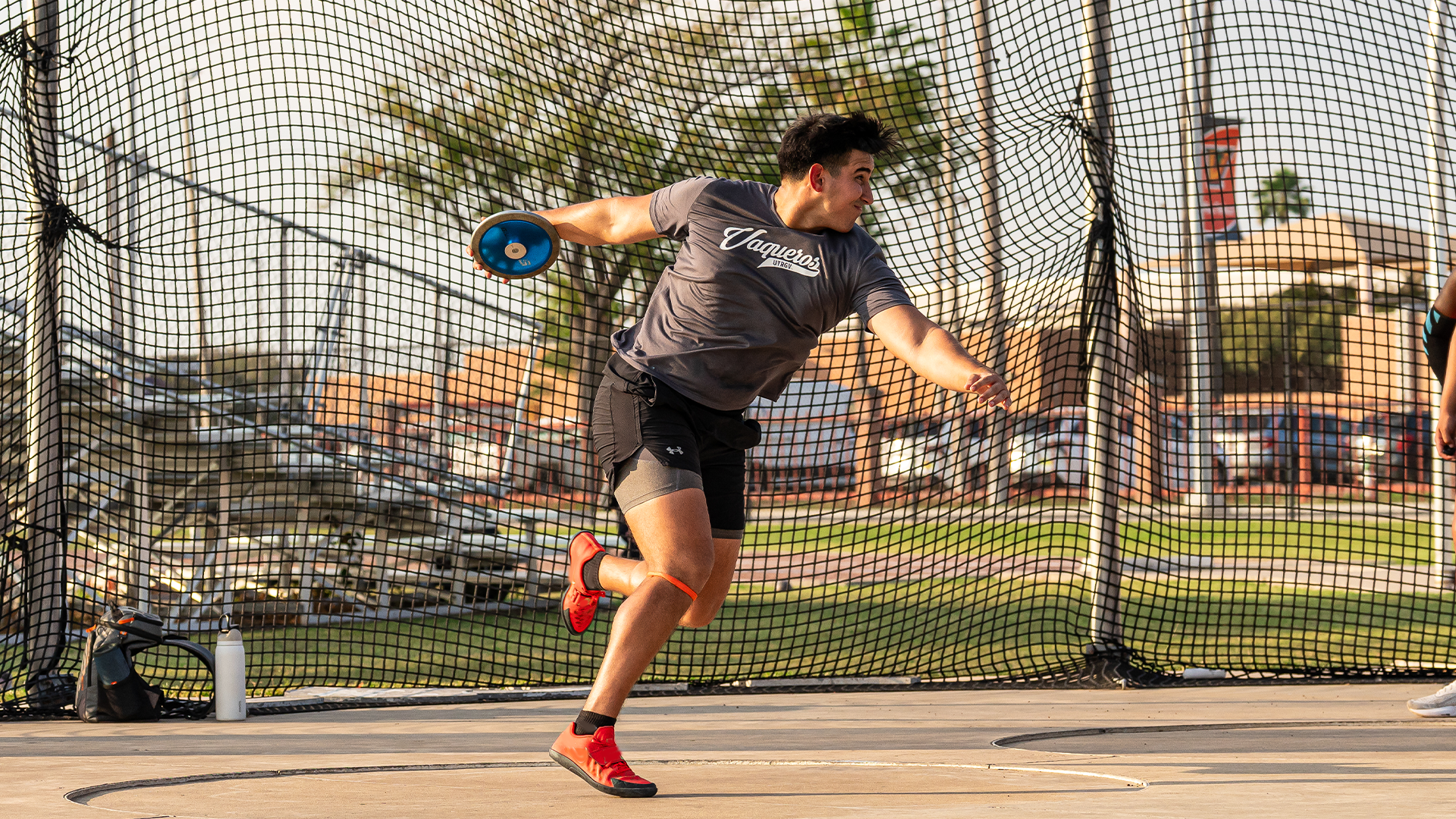 Photo is of UTRGV track & field senior Tristan Solis wearing a gray shirt with Vaqueros written in script across the chest, gray shorts and red shoes throwing discus in the throwing ring at the UTRGV Soccer and Track & Field Complex