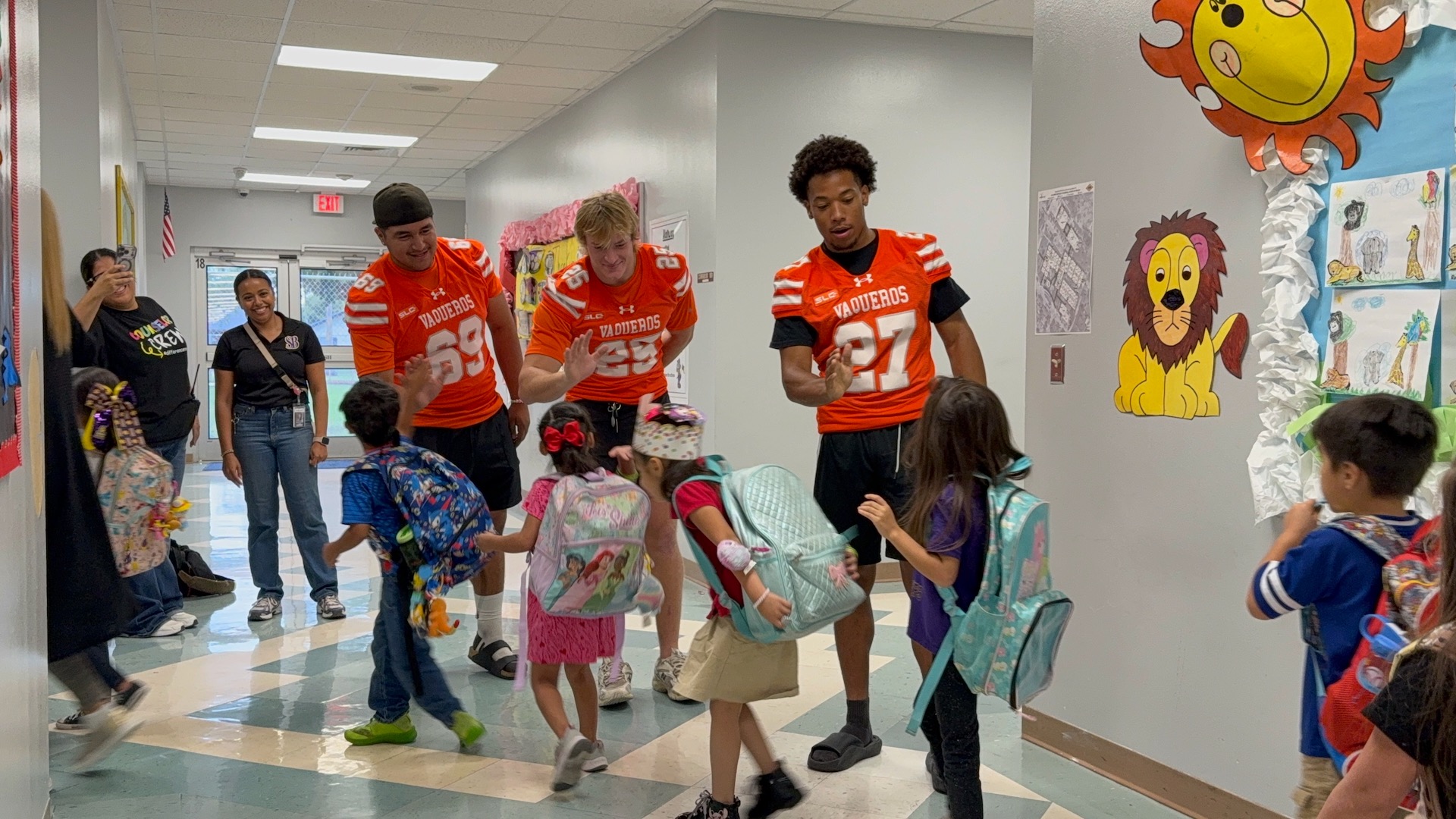 Image shows UTRGV football players Agustin Renteria, Walker Diharce and Andrew Earls in their orange jerseys giving high fives to students as they walk down a hallway at a local elementary school